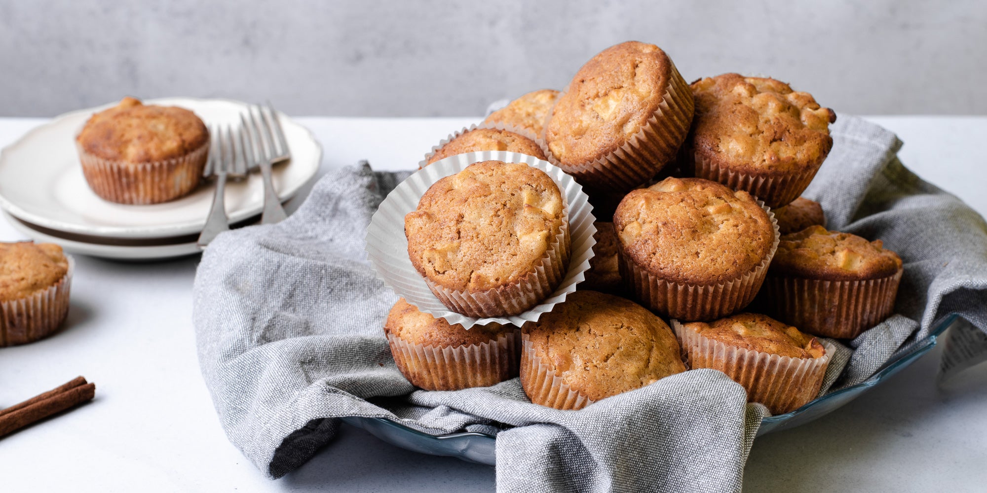 A pile of Calorie Conscious Apple & Cinnamon Muffins on a grey cloth, with a muffin on a plate with forks in the background