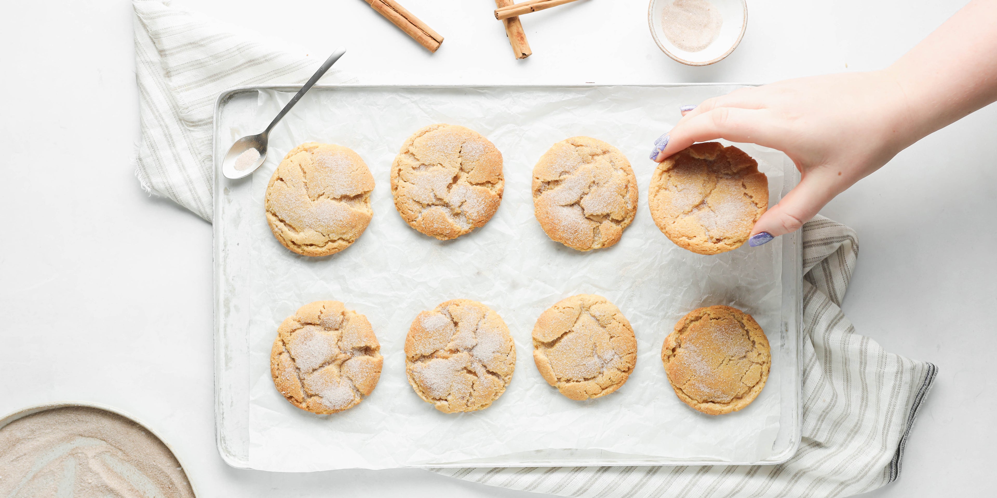 Top view of a batch of Churro Cookies on a baking tray, with a hand reaching for a cookie - sprinkled with sugar