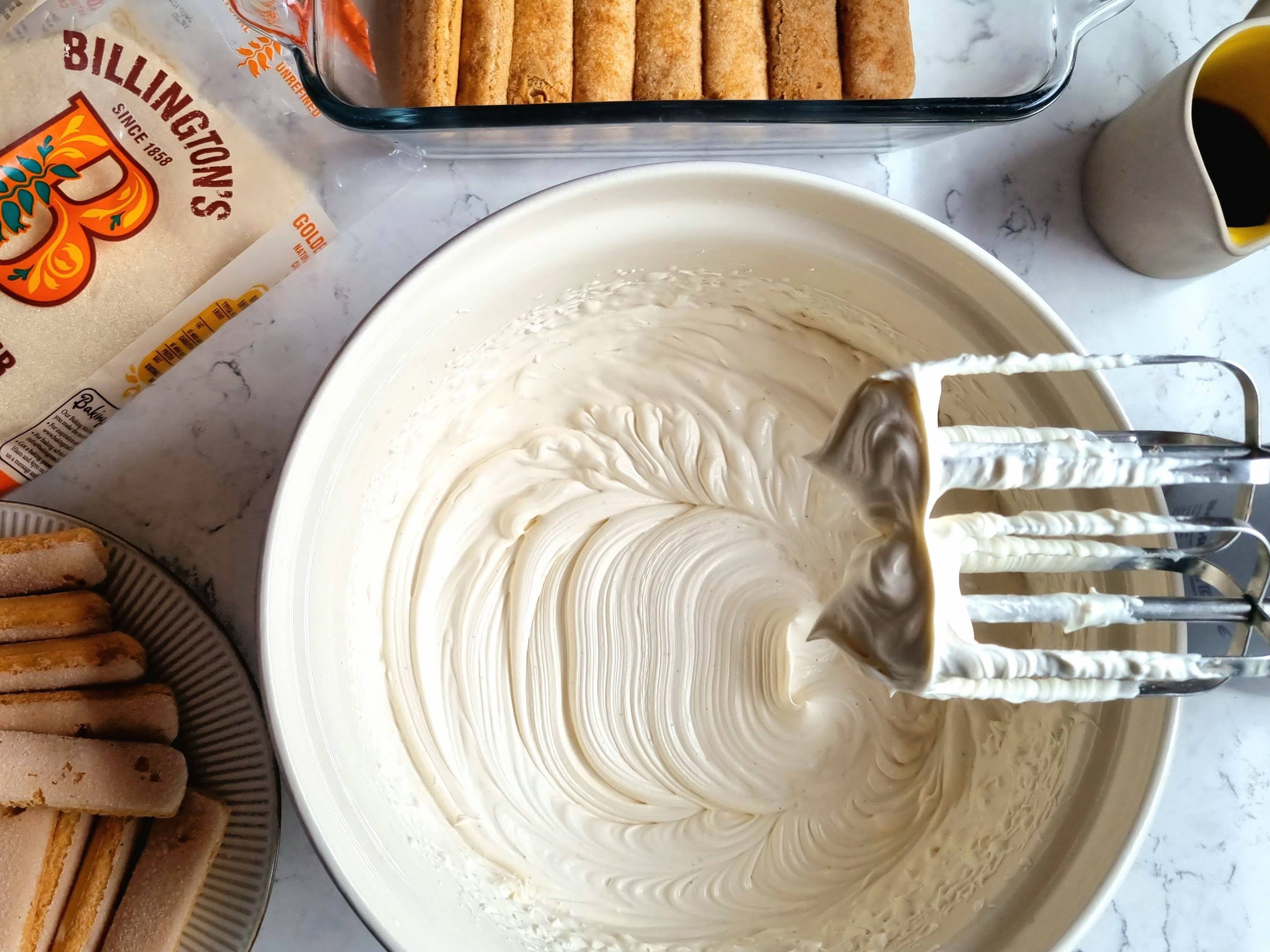 Bowl of whipped cream for tiramisu, with a small jug of coffee, baking tray and plate with sponge fingers and pack of golden caster sugar