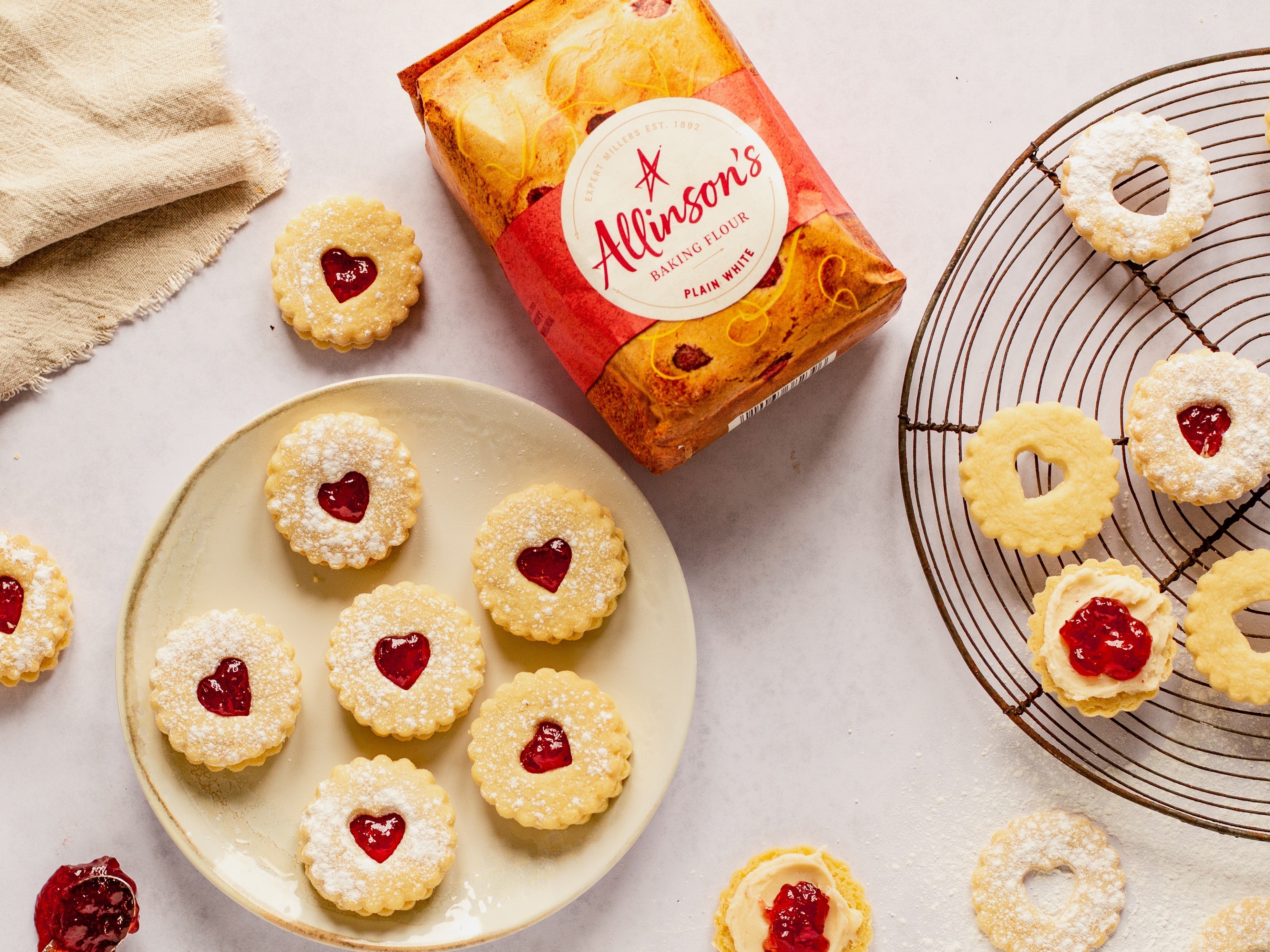 Plate of jam dodger biscuits. Biscuits half assembled on cooling rack next to plate. Flour pack in shot