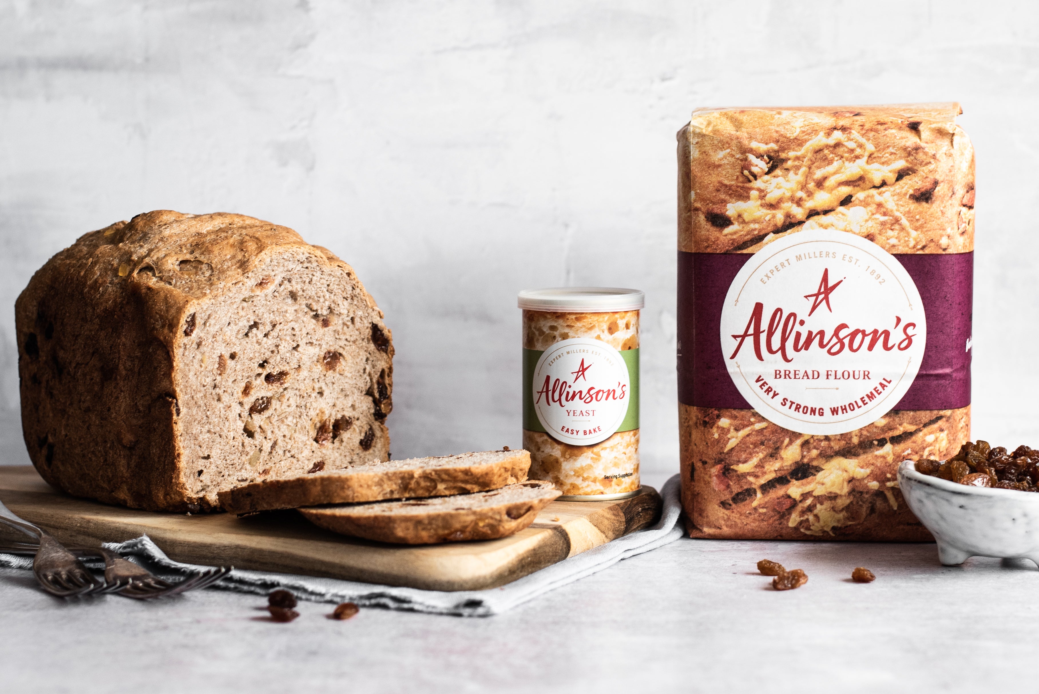 Fruit bread with slices cut infront. Two forks. Tin of yeast. Flour pack. Bowl of sultanas