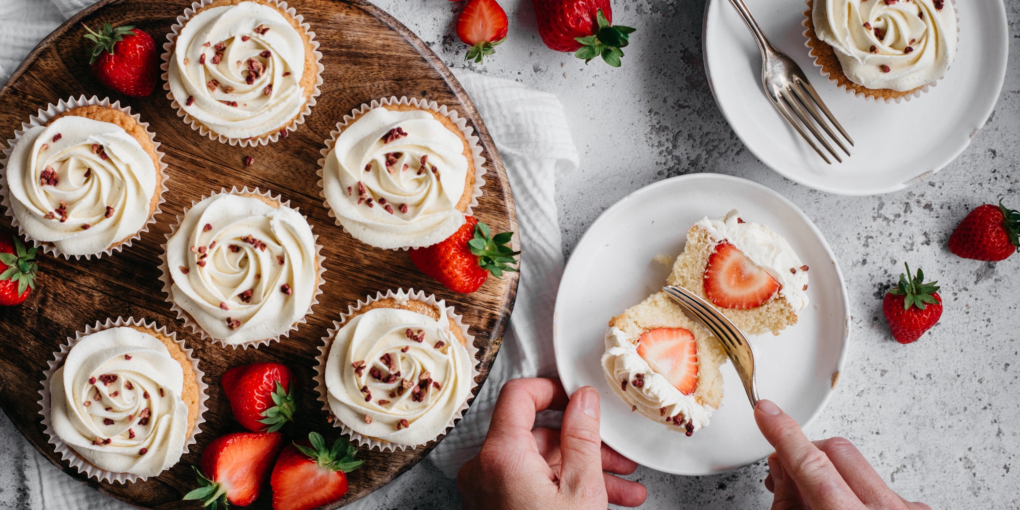 Top down shot of a circle plate of cupcakes and strawberries. A white plate on the right side with a cupcake cut in half and a hand holding a fork