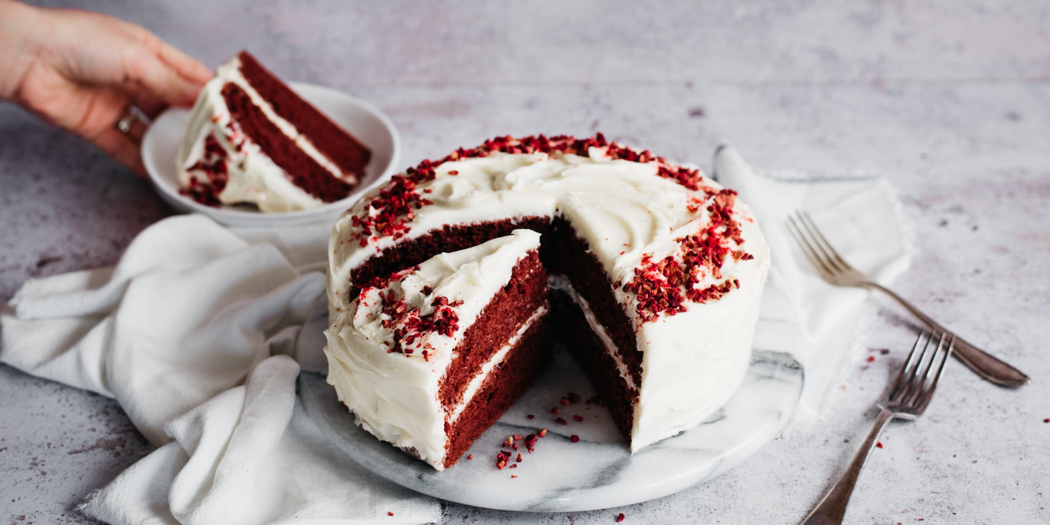 Red Velvet Cake with slices cut out of it showing the rich, red insides. Hand holding a plate with a slice of Red Velvet Cake
