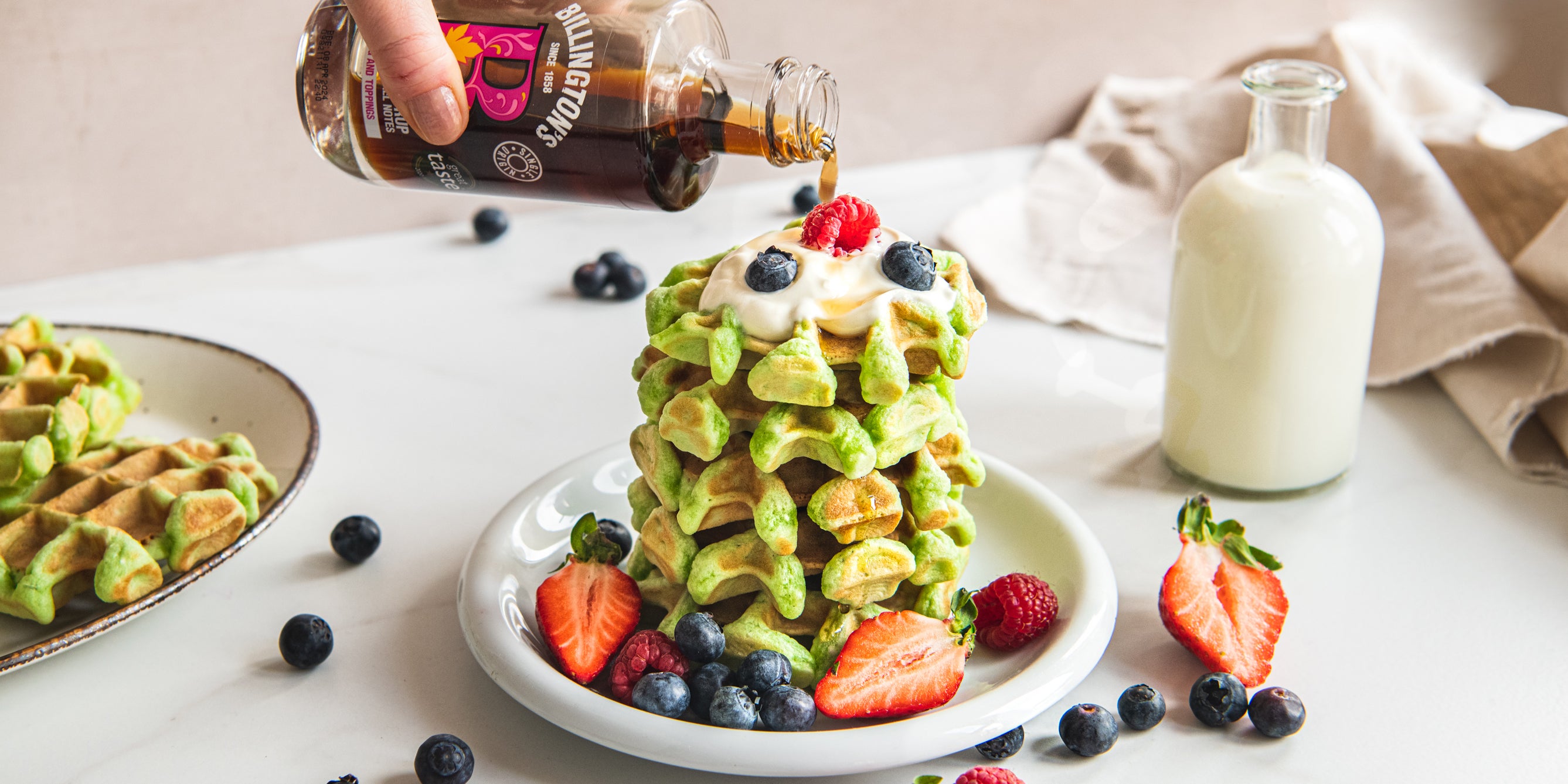 A fresh stack of green Pandan Waffles sprinkled with berries and being drizzled with Billington's maple syrup