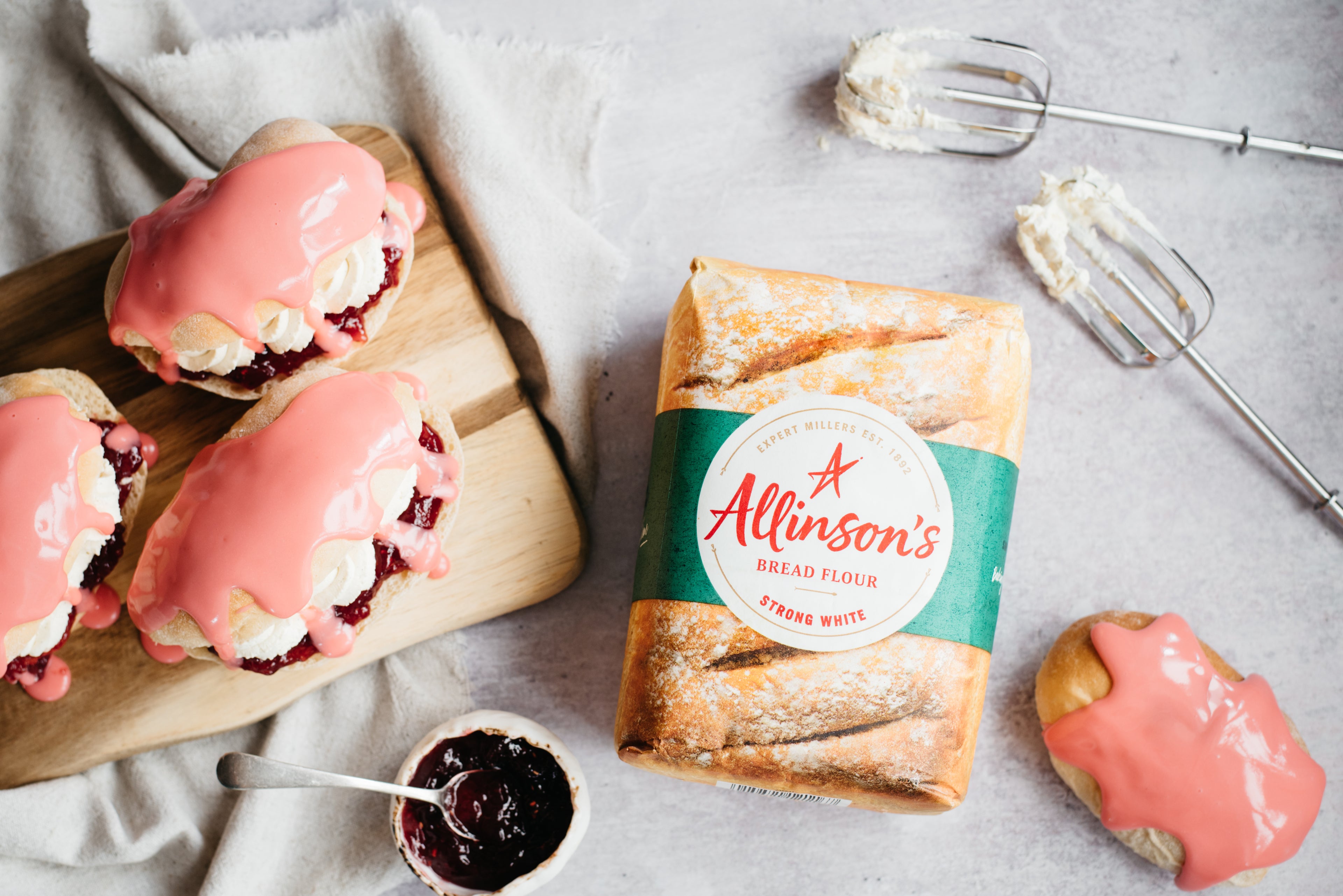 Flat lay of Iced Finger Buns drizzled in pink icing, next to a bag of Allinson's strong white flour, and whisks covered in mixture