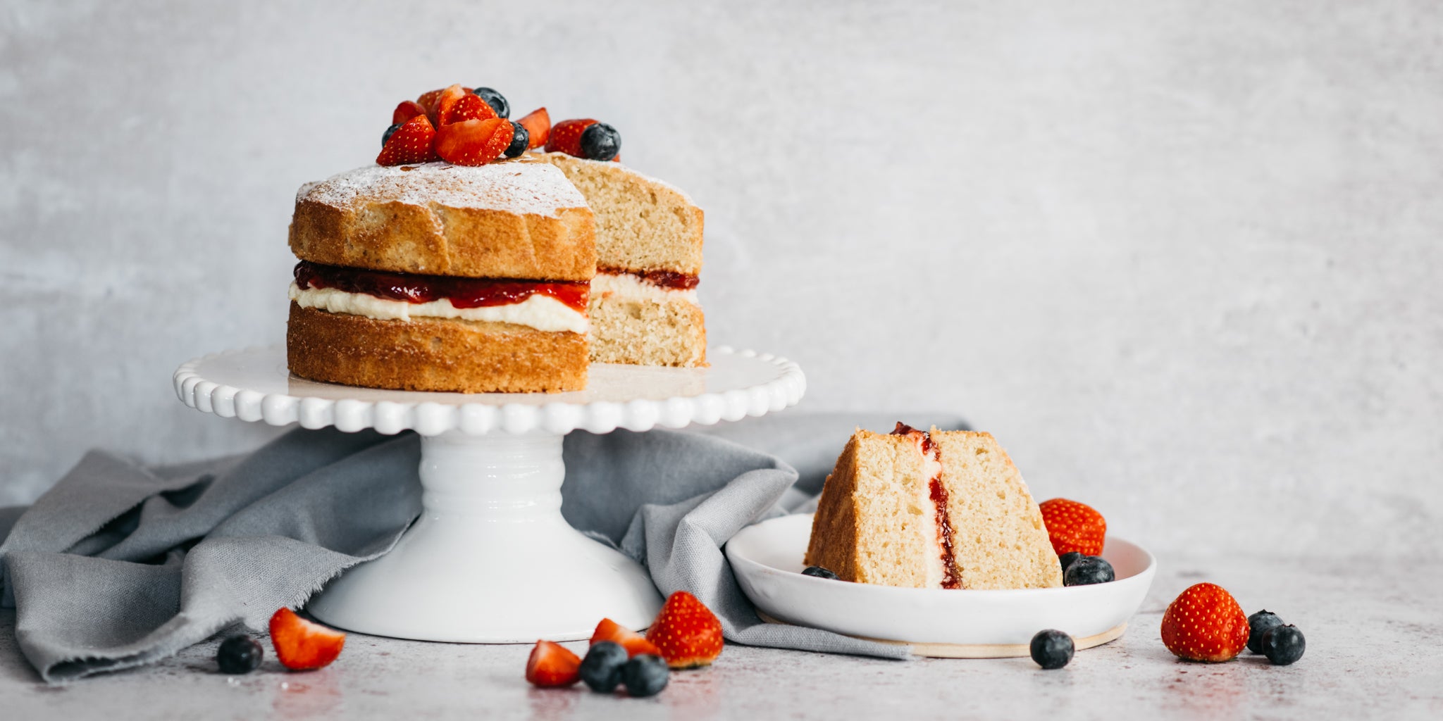 Wholemeal Victoria Sponge on a cake stand, with a slice cut out of it served on a plate and garnished with fresh summer berries