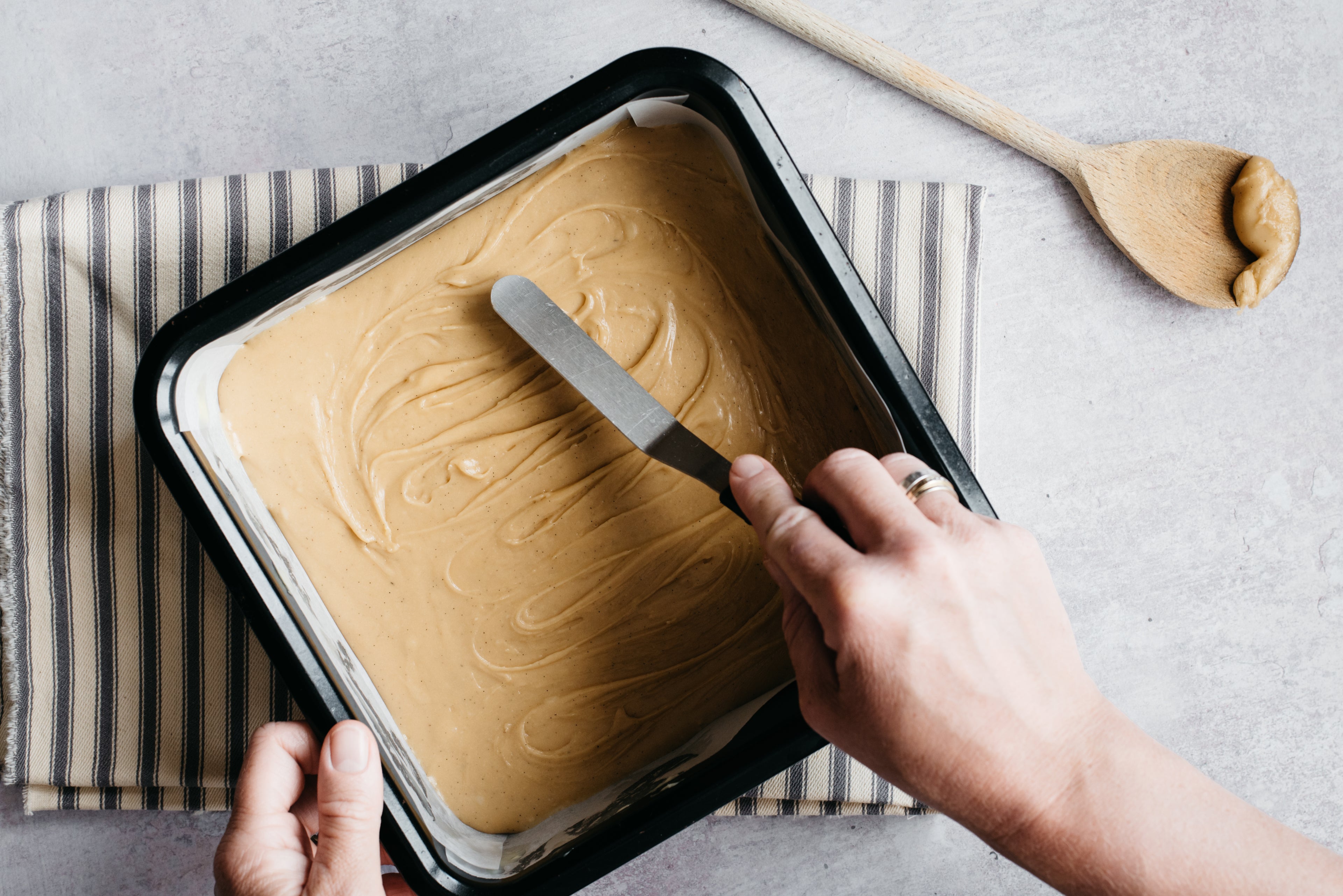 Hand smoothing down the fudge with a palette knife in a square baking tin. Stripe towel underneath and a wooden spoon with mixture on at the side