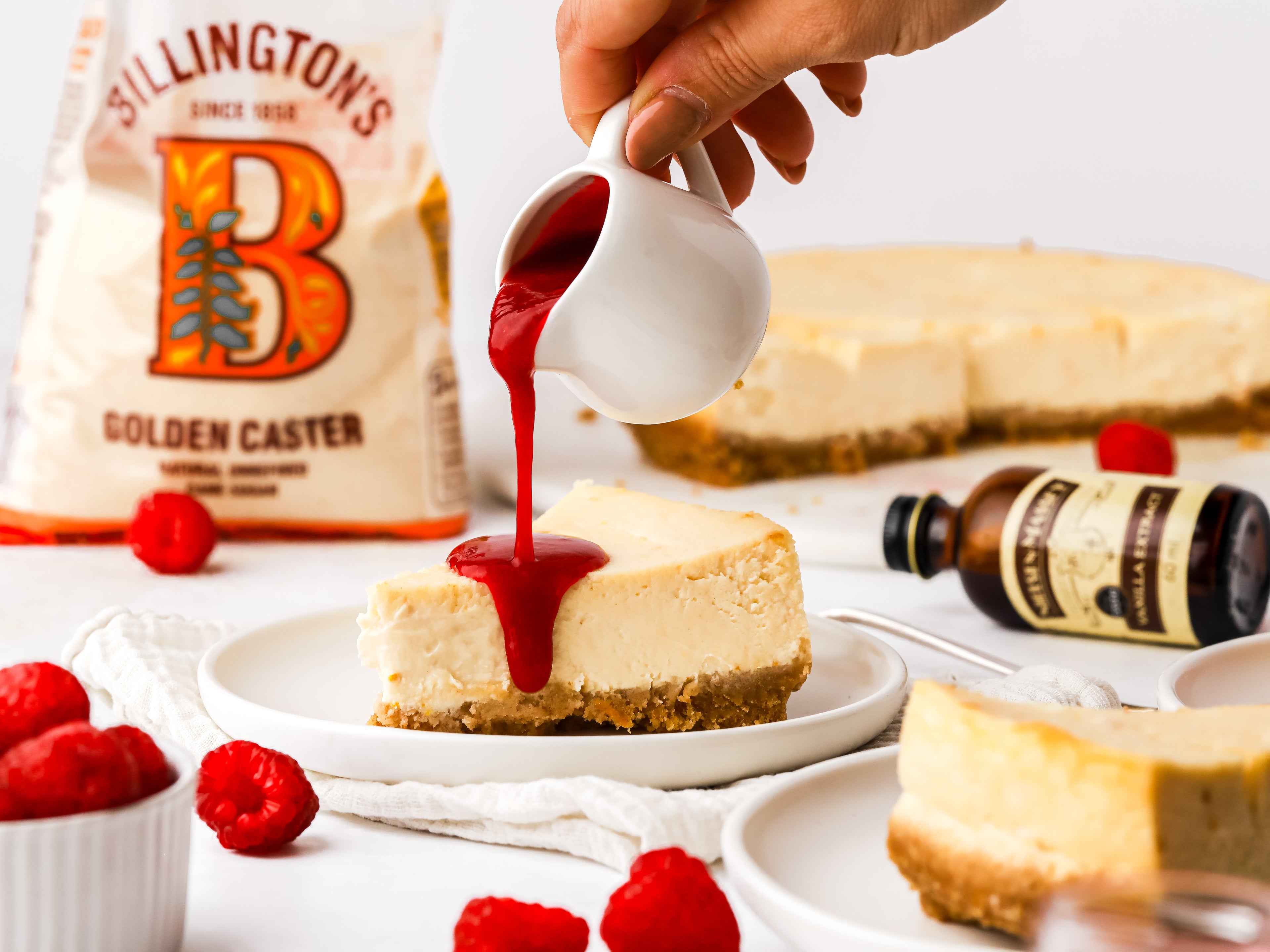 Raspberry sauce being poured on top of a slice of baked cheesecake with sugar pack in background and vanilla bottle on its side