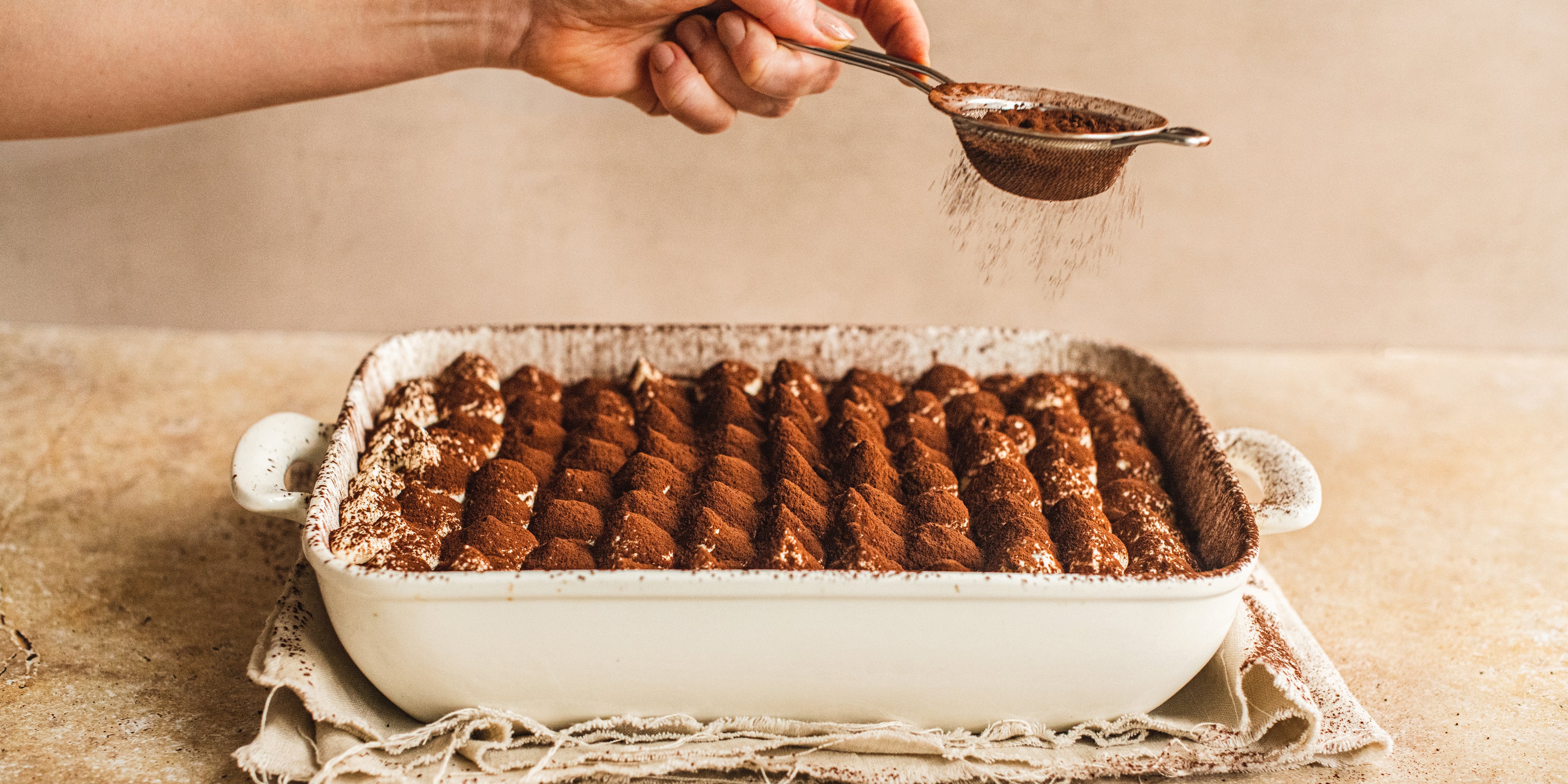 Tray of Vegan Tiramisu being hand sprinkled with cocoa powder to finish the dessert off