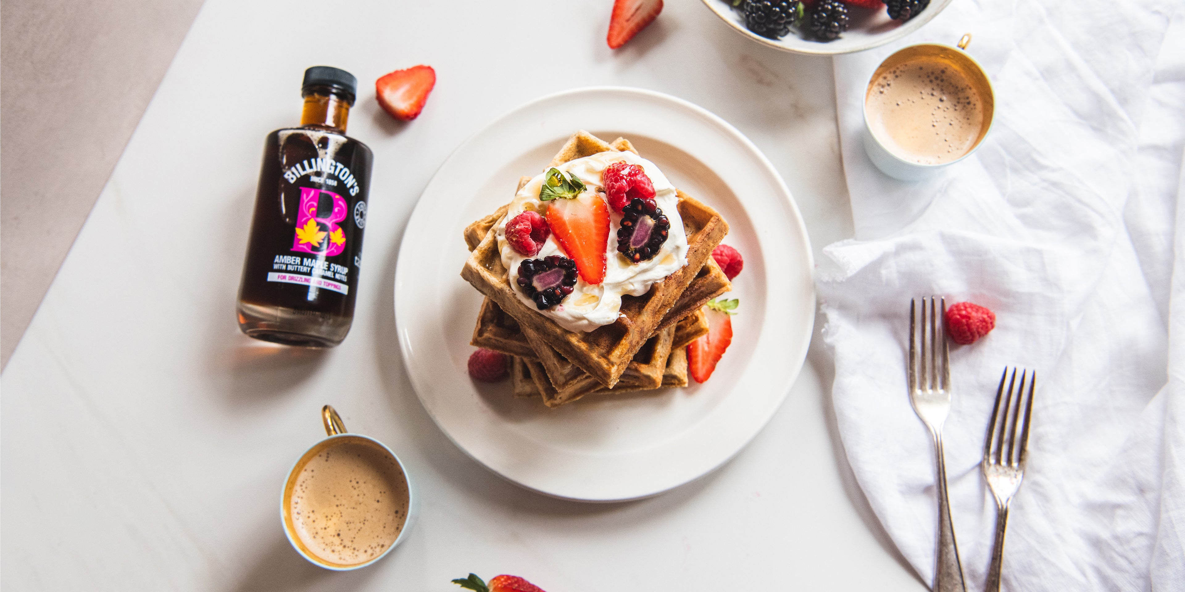 Top view of Wholemeal Banana Waffles next to a bottle of Billington's Syrup, berries and cutlery 