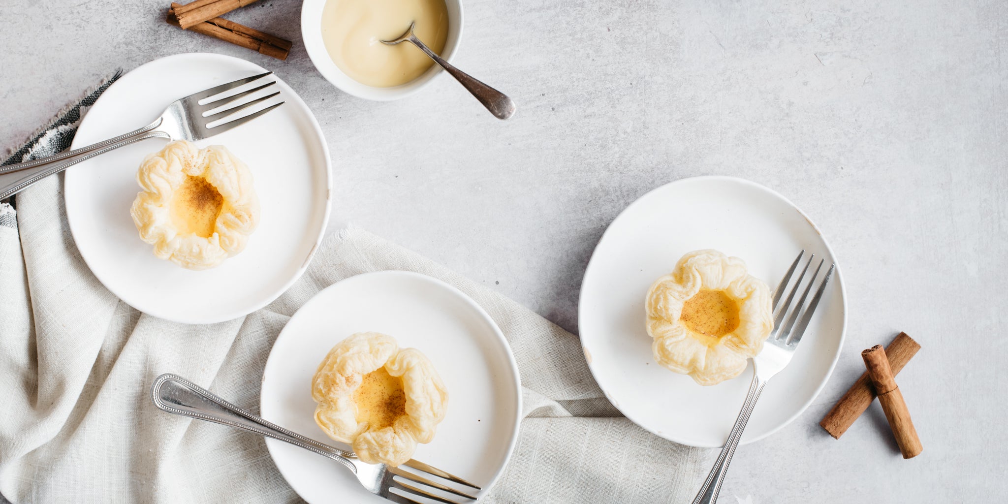 3 small white plates with Portuguese Custard Tarts on and forks and cinnamon sticks beside