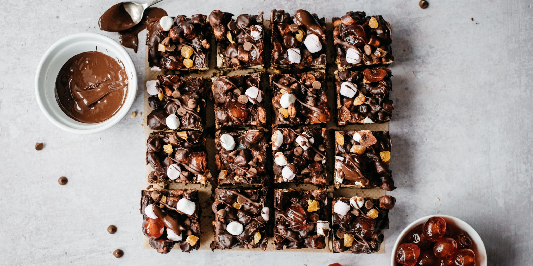 Top view of Vegan Rocky Road traybake sliced into squares, next to a bowl of melted chocolate and glace cherries