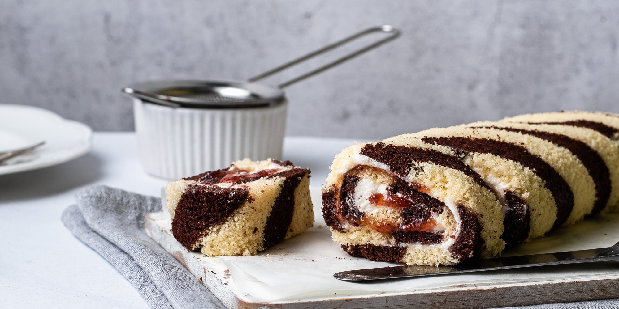 Close up of Chocolate & Vanilla Swiss Roll served on baking paper and linen, with a slice of Swiss Roll lay next to it. 
