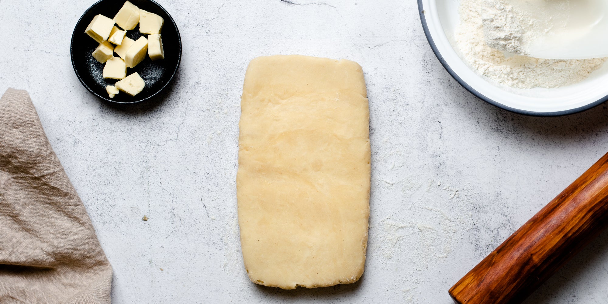 Puff Pastry rolled out onto a surface next to a dish of butter, bowl of flour and rolling pin