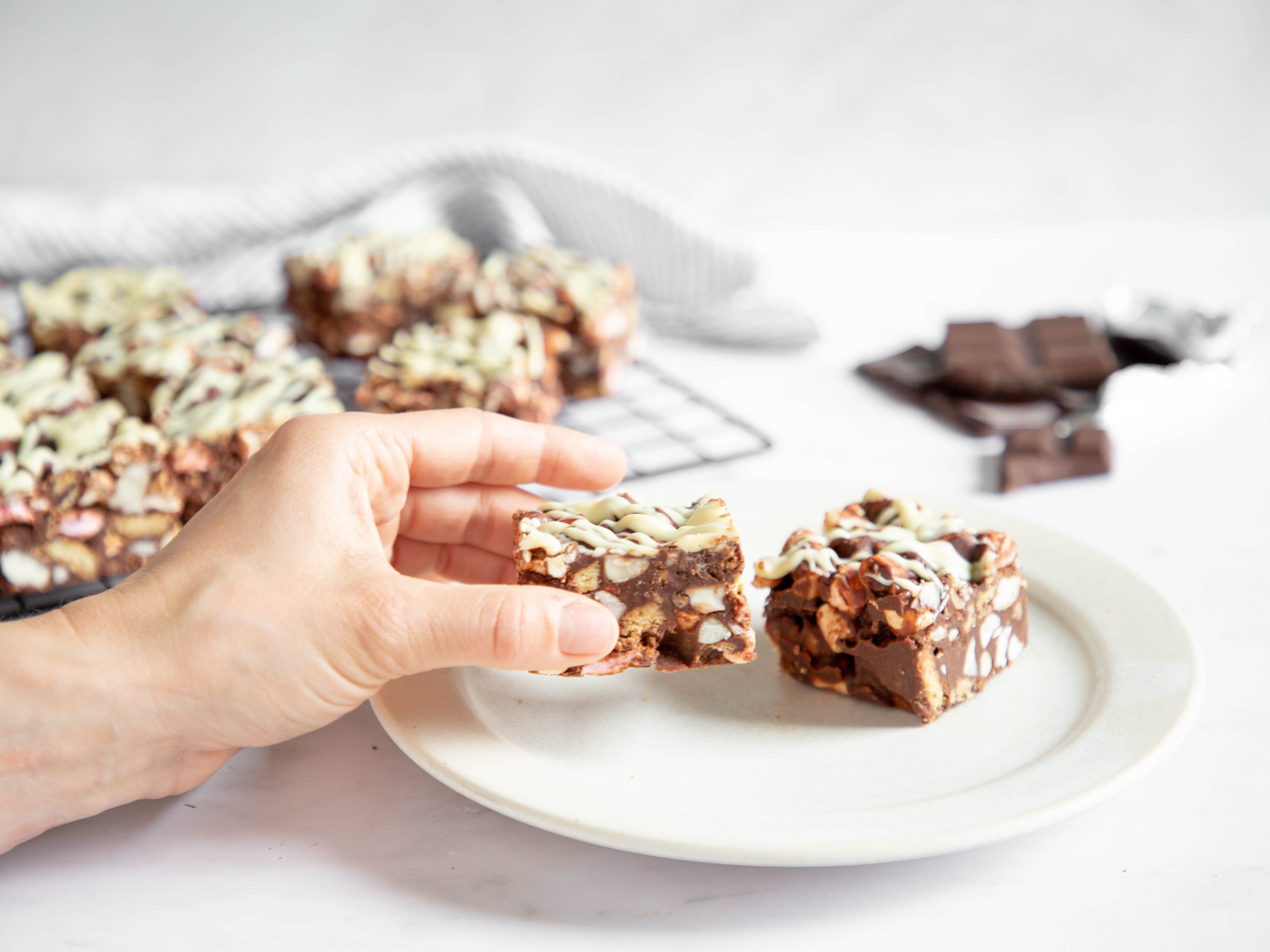 Hand reaching in to take a slice of rocky road from a white plate. More squares of rocky road on a cooling rack in the background along with some chocolate chunks