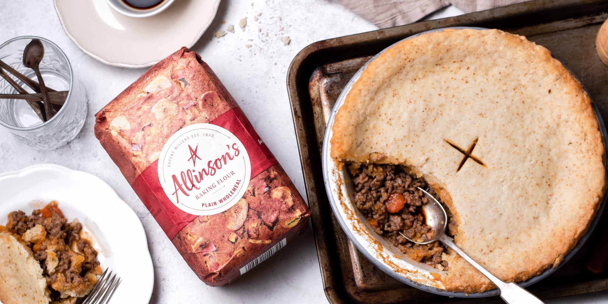 Suet Crusted Beef & Onion Pie on a baking tray, with a spoon serving some of the pie filling. Next to a bag of Allinson's Plain Wholemeal flour and a plate with a serving of pie.