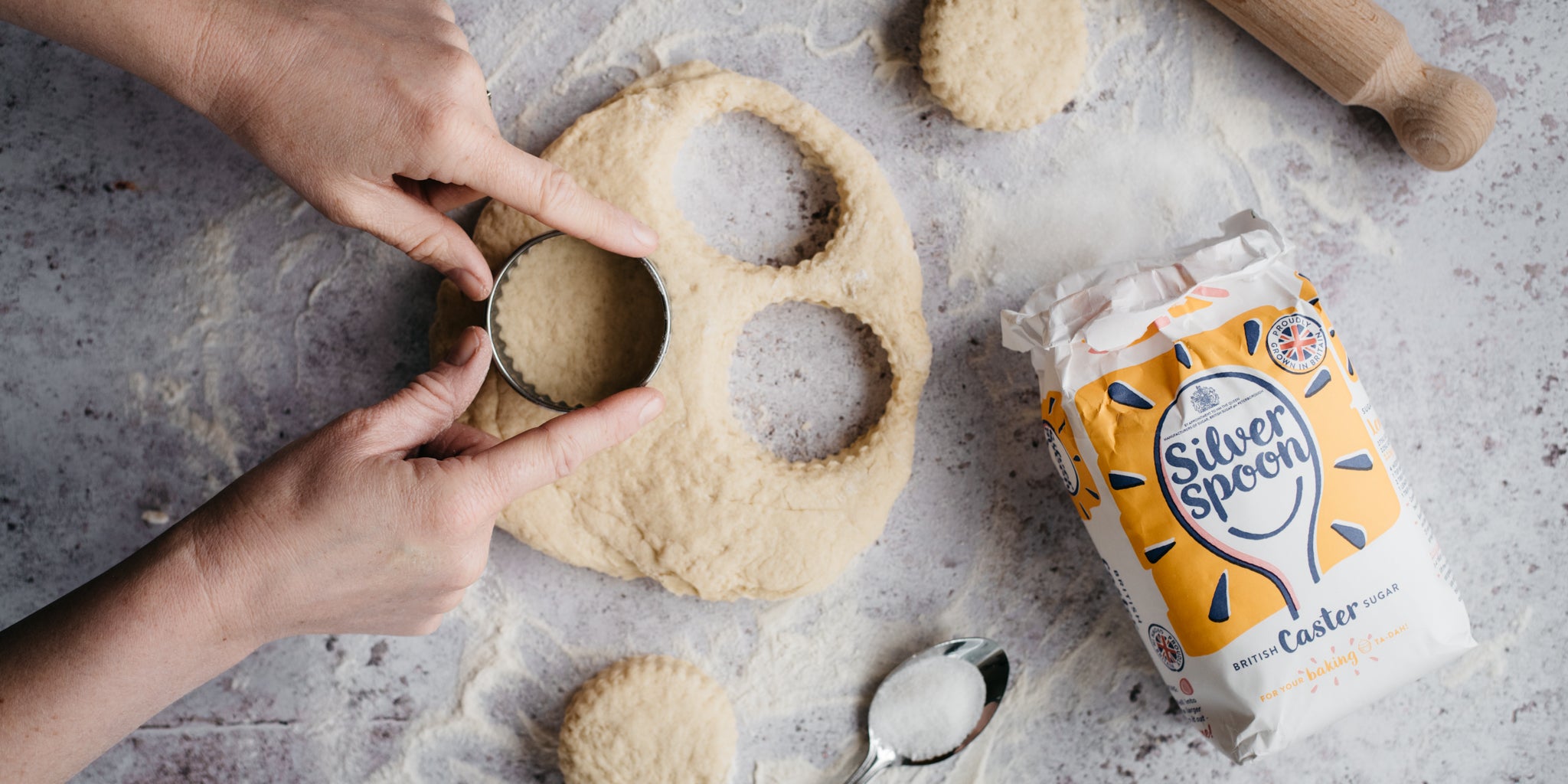 Top view of Vegan Scones being cut out of vegan dough with hands holding a cutter, next to a flat lay bag of Silver Spoon Caster Sugar