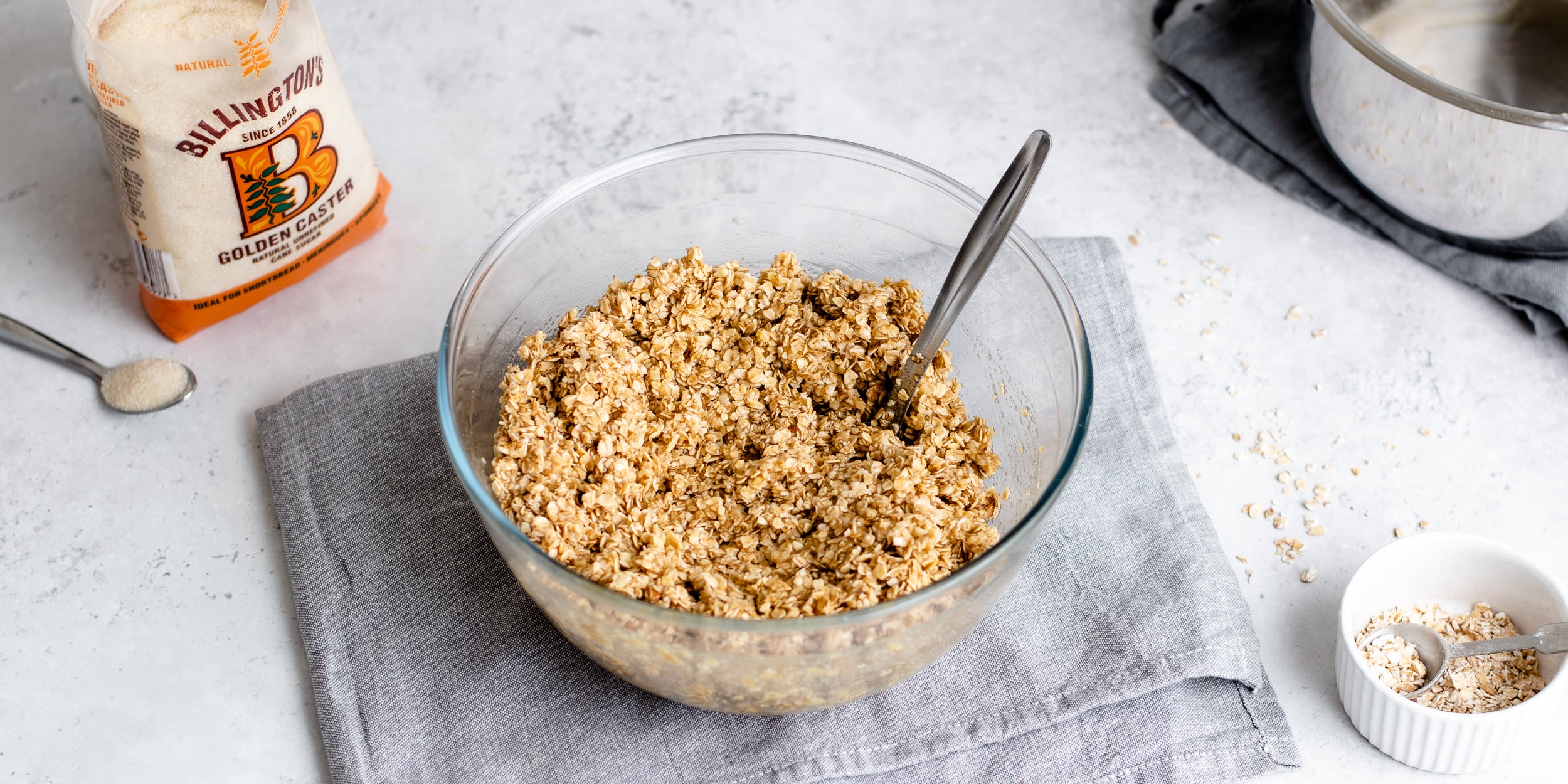 Easy Flapjack mix in a glass bowl being combined. Next to a bag of Billington's Golden Caster Sugar