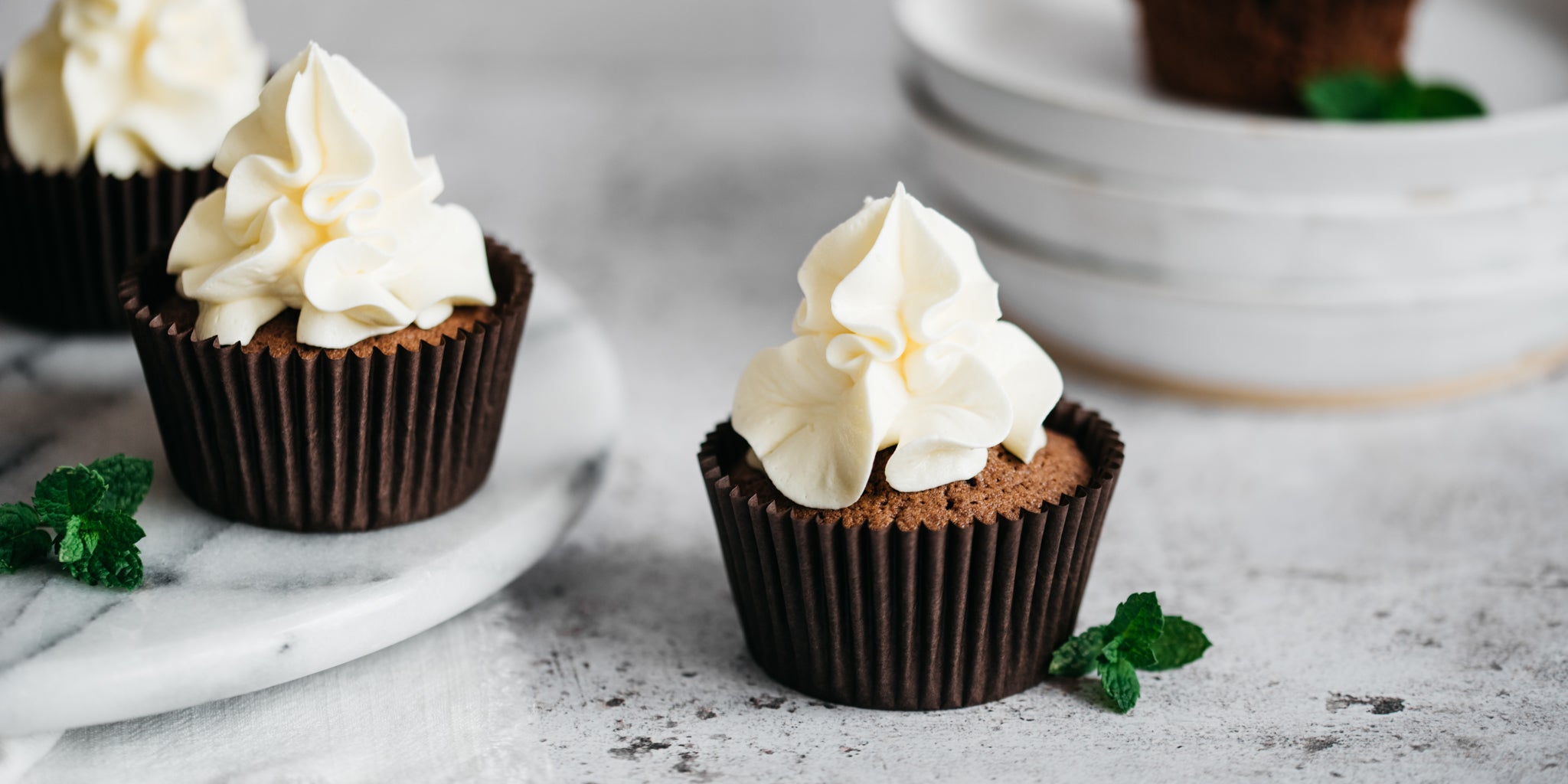 Close up of a Chocolate & Peppermint Cupcake next to a mint leaf garnish, and topped with fluffy peppermint buttercream