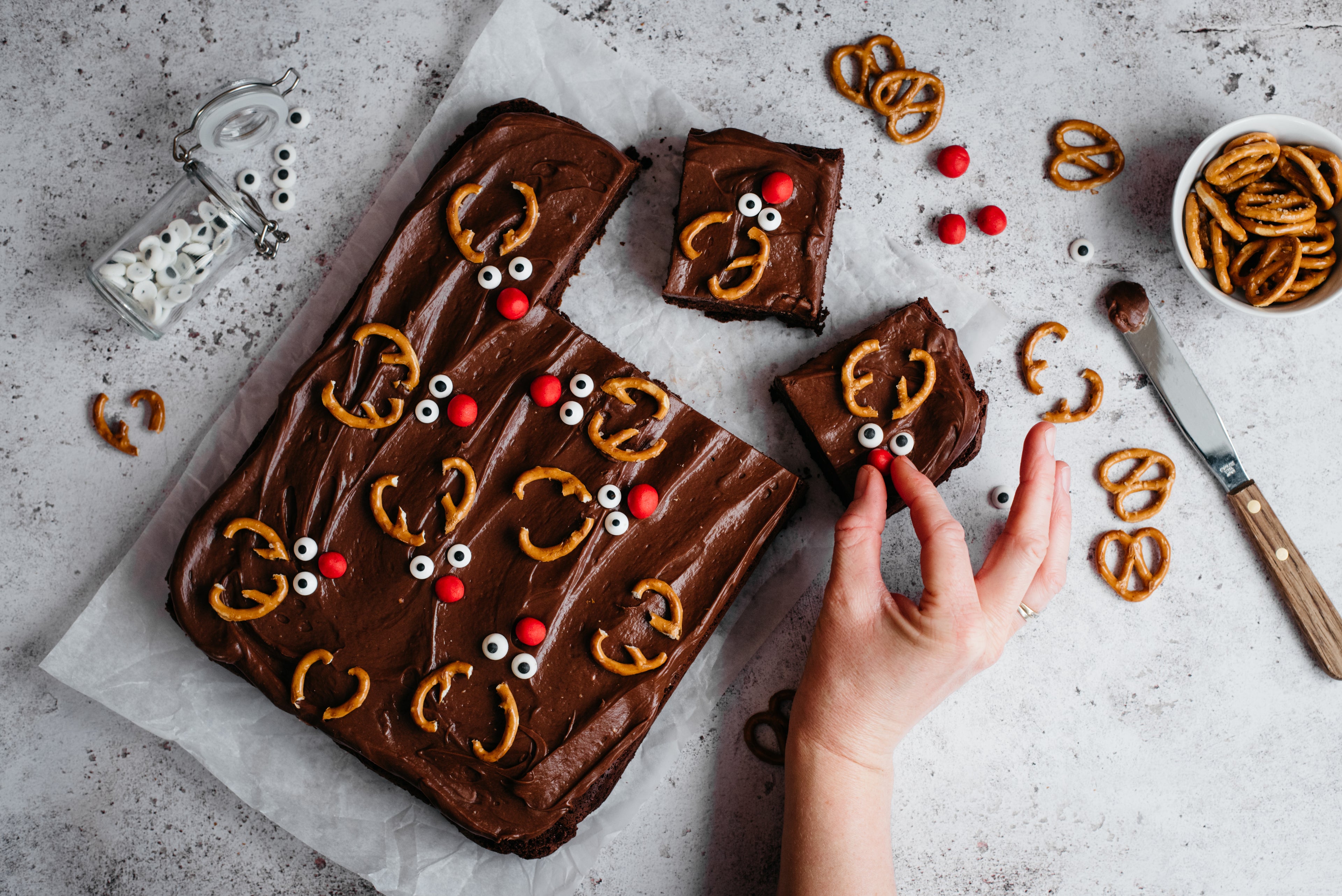 Top view of Chocolate & Caramel Reindeer Traybake with a hand reaching to decorate with red sweets. Next to a bowl of pretzels