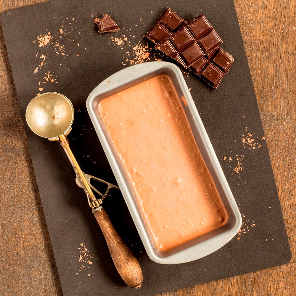 Tub of homemade chocolate ice cream next to squares of chocolate and a silver ice cream scoop