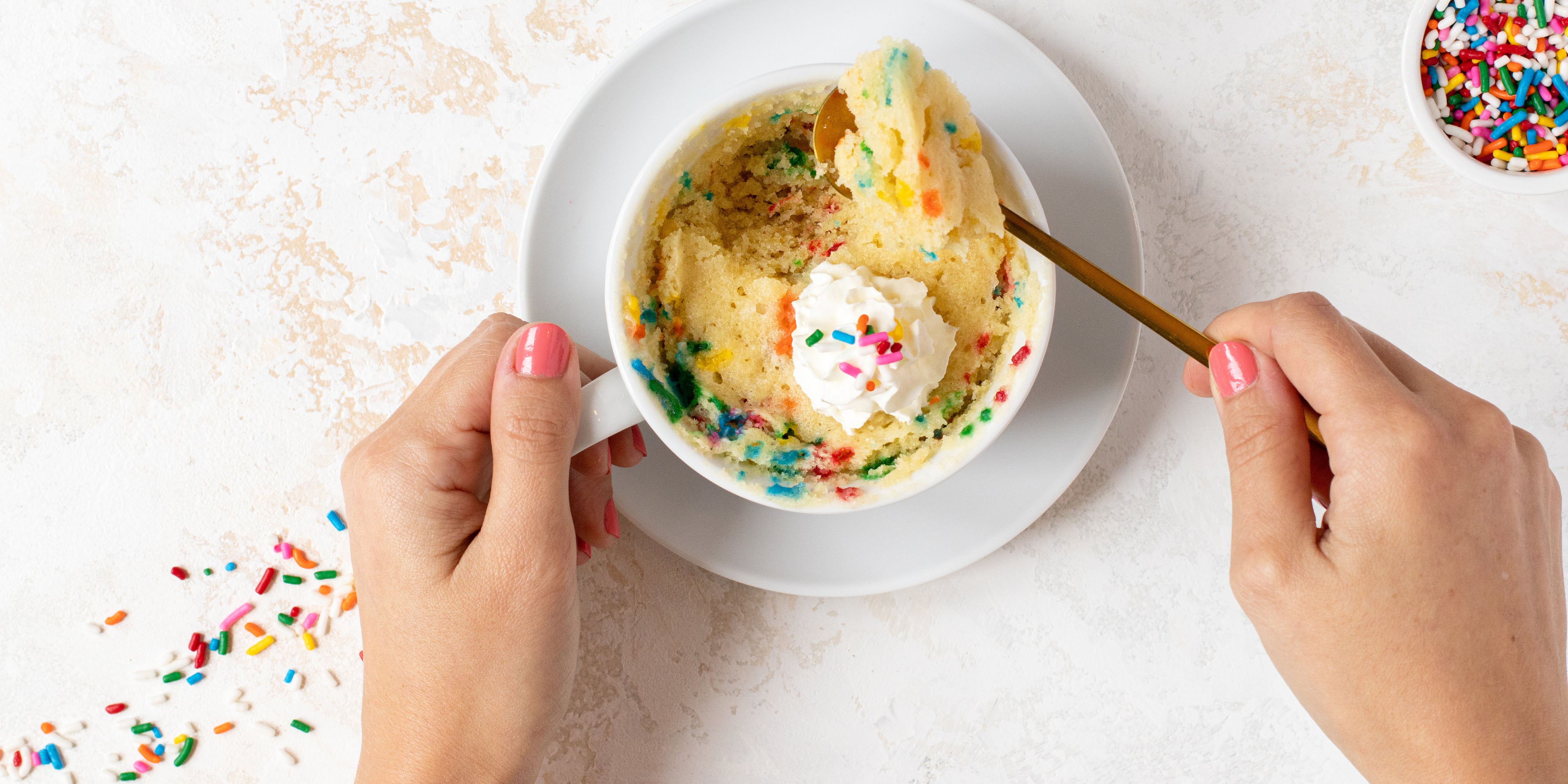 Top view of Birthday Cake Mug Cake with a hand dipping into a spoon and sprinkles next to it 