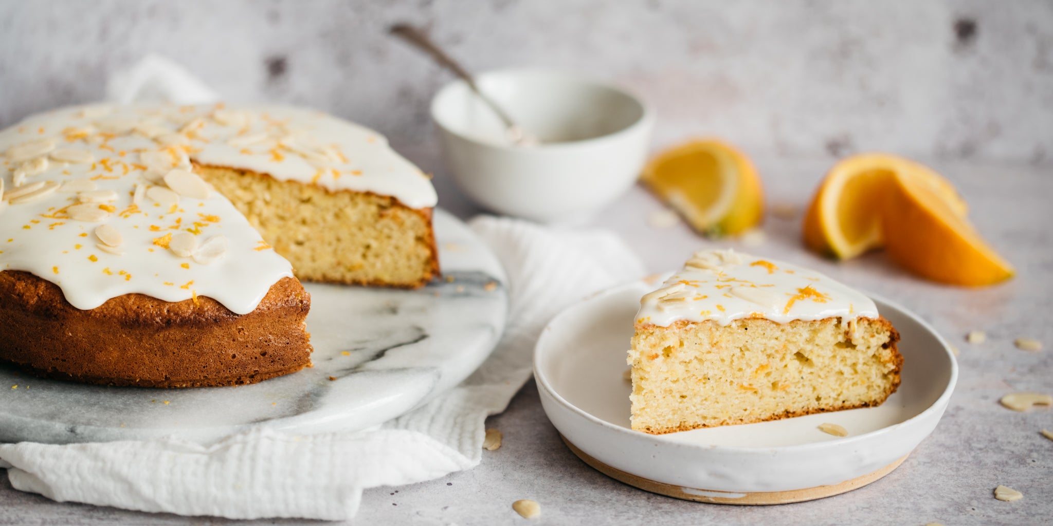 Cake topped with icing and a slice removed on a plate infront. Orange slices in the background