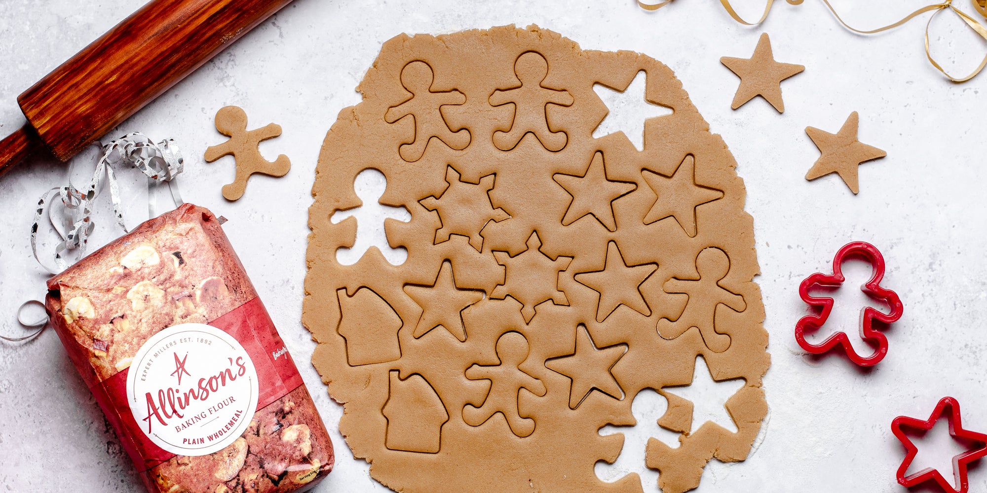 Top view of rolled out Gingerbread Dough, with shapes cut out of it, next to cookie cutters and a bag of Allinson's plain flour