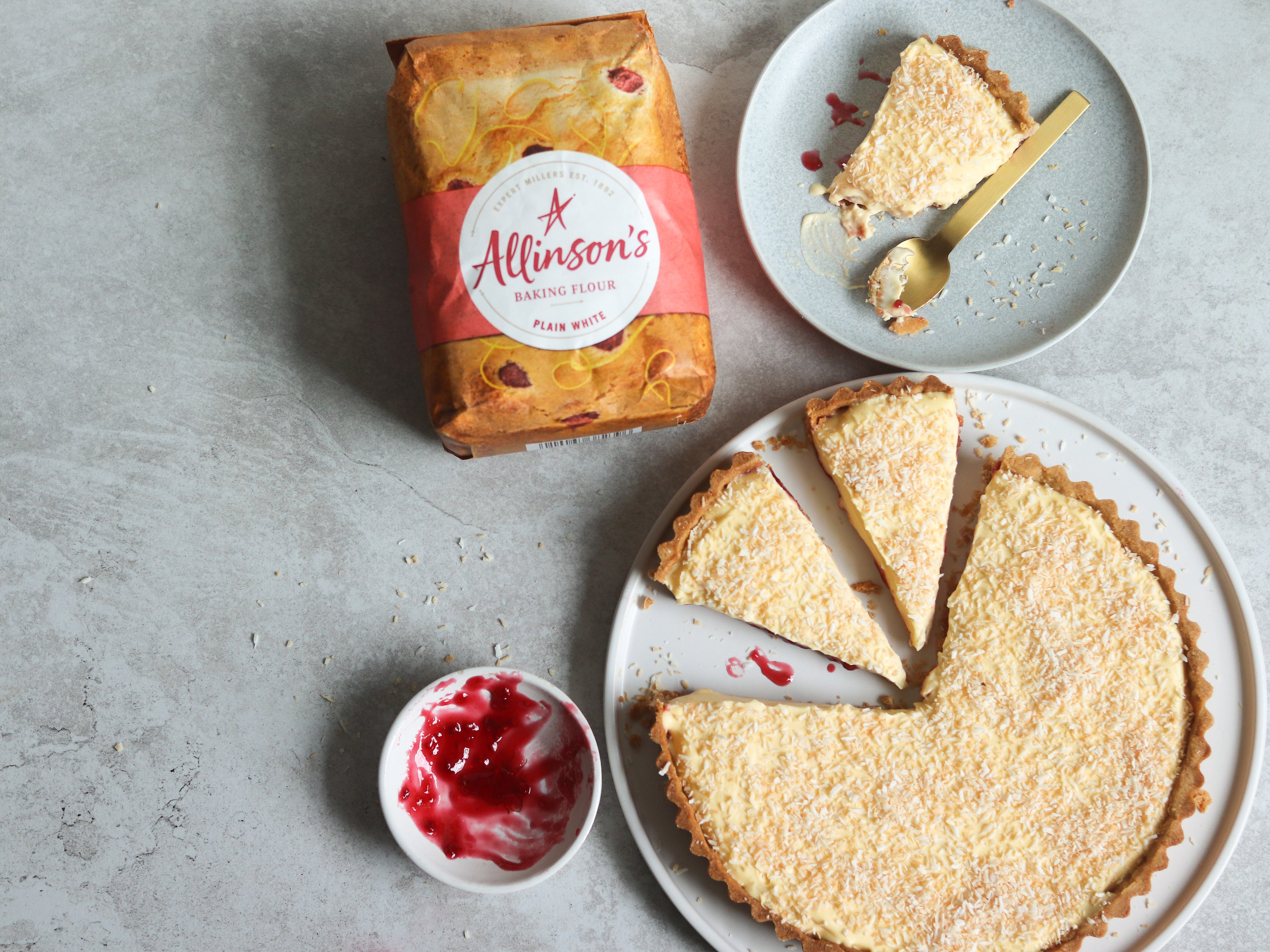 Top view of a Manchester Tart sliced with pieces to serve. Next to a bag of Allinson's Plain Flour and a slice of Manchester Tart on a plate with a spoon covered in toasted coconut