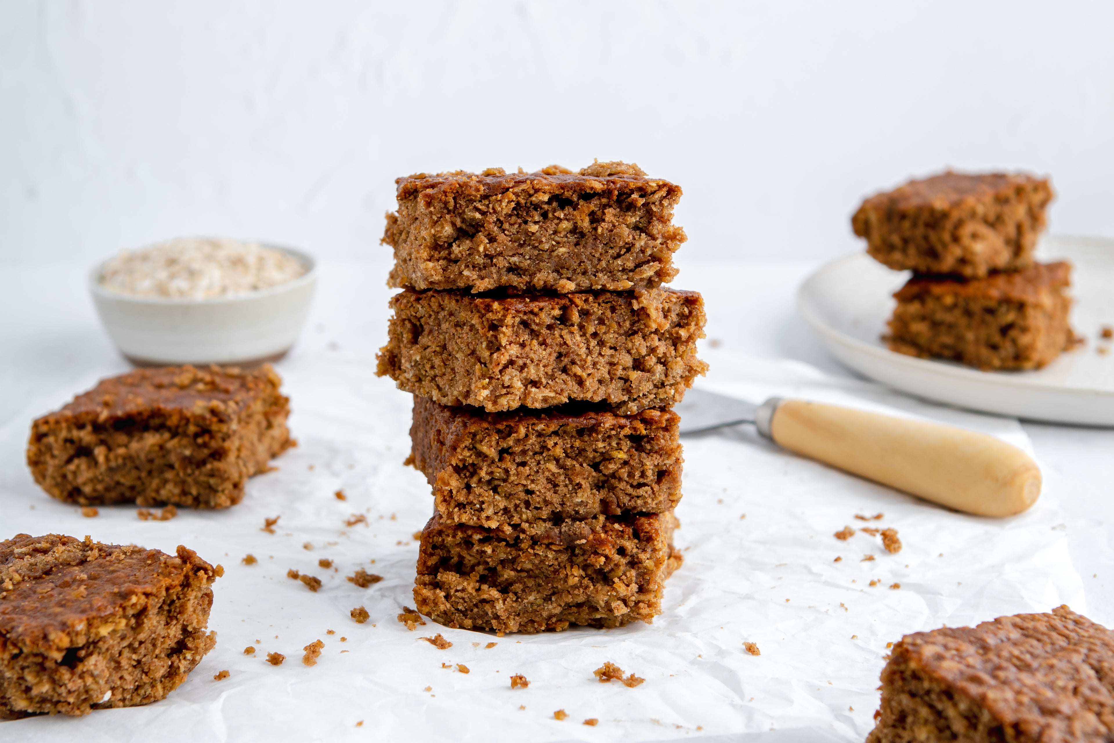 Squares of Sticky Yorkshire Parkin stacked on top of each other, surrounded by crumbs with a bowl of oats in the background
