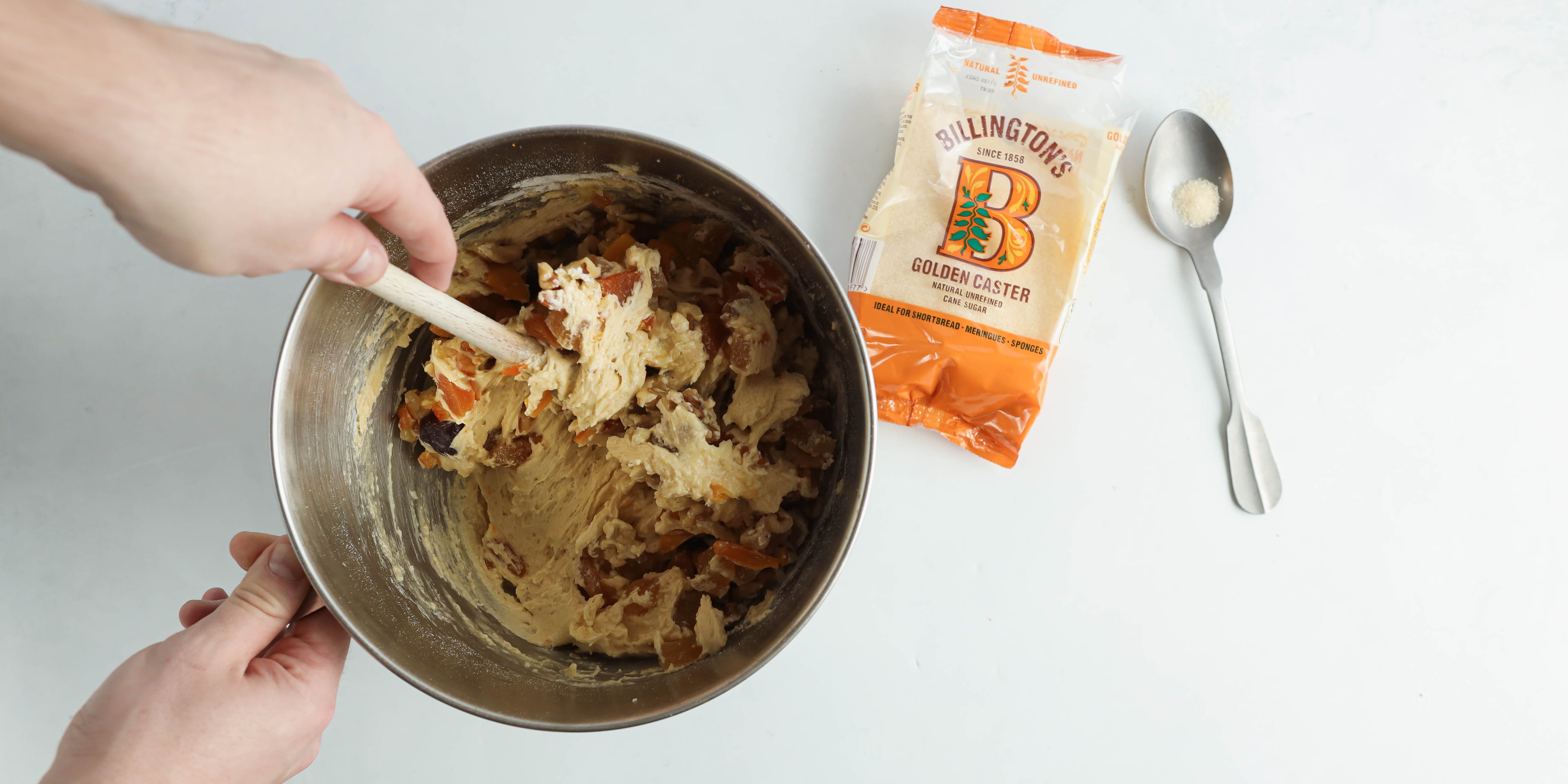 Top view of a hand mixing the Fruit Cake mixture, next to a flat lay bag of Billington's Golden Caster Sugar