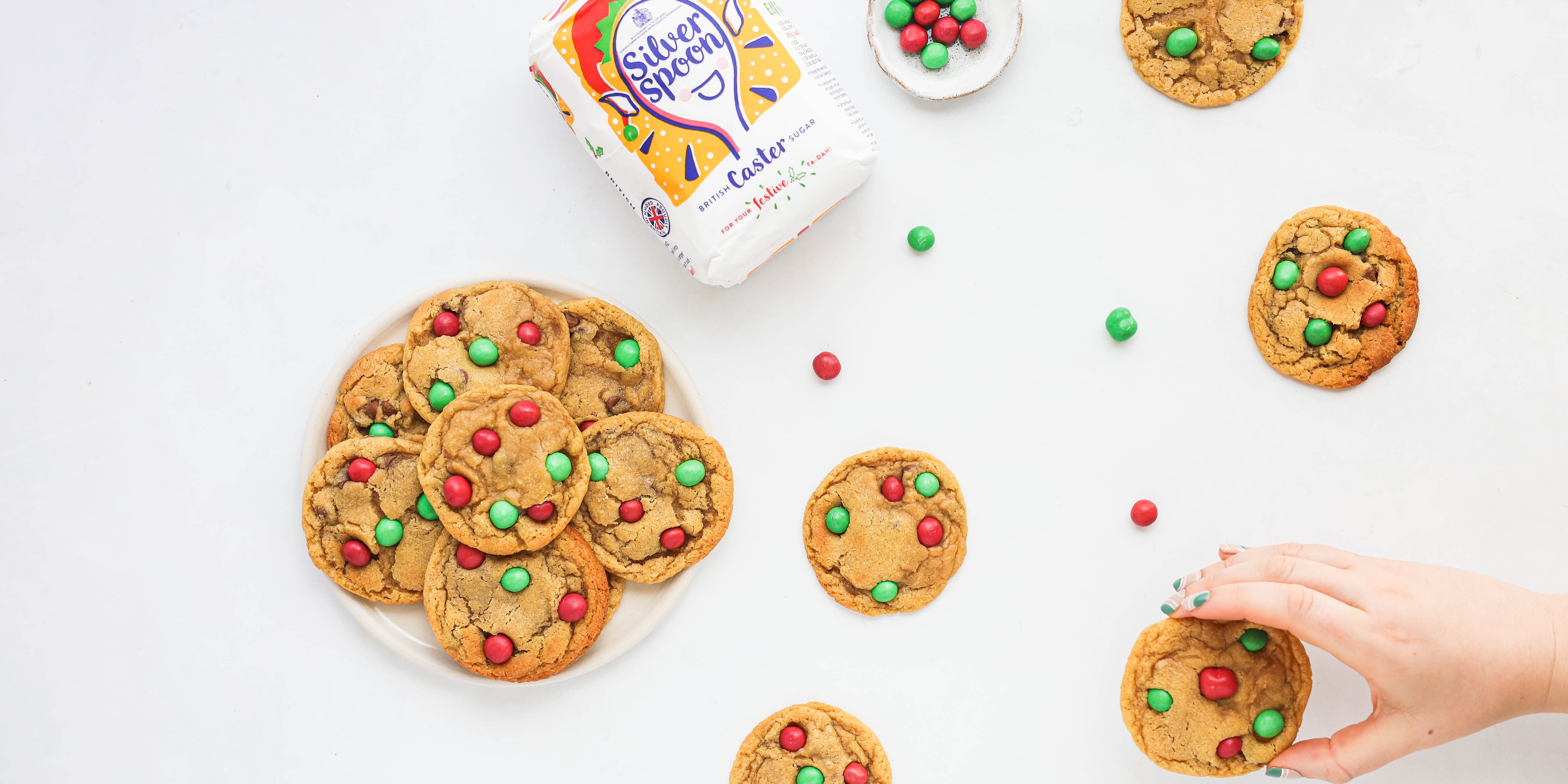 Top view of Copycat Millies Cookies with red and green m&m's for Christmas. Hand reaching for a cookie next to a bag of Silver Spoon caster sugar