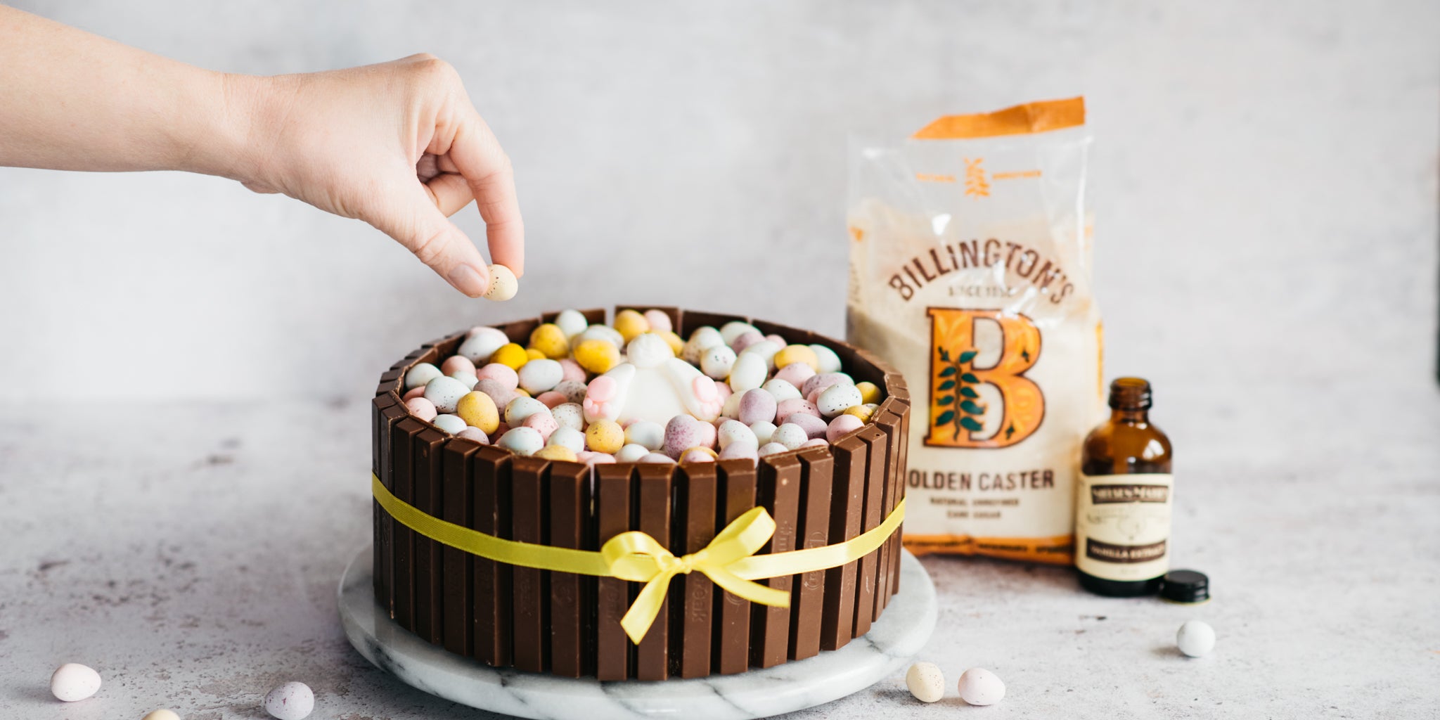 Cake decorated with kitkats, yellow ribbon and a hand reaching in adding decoration on top