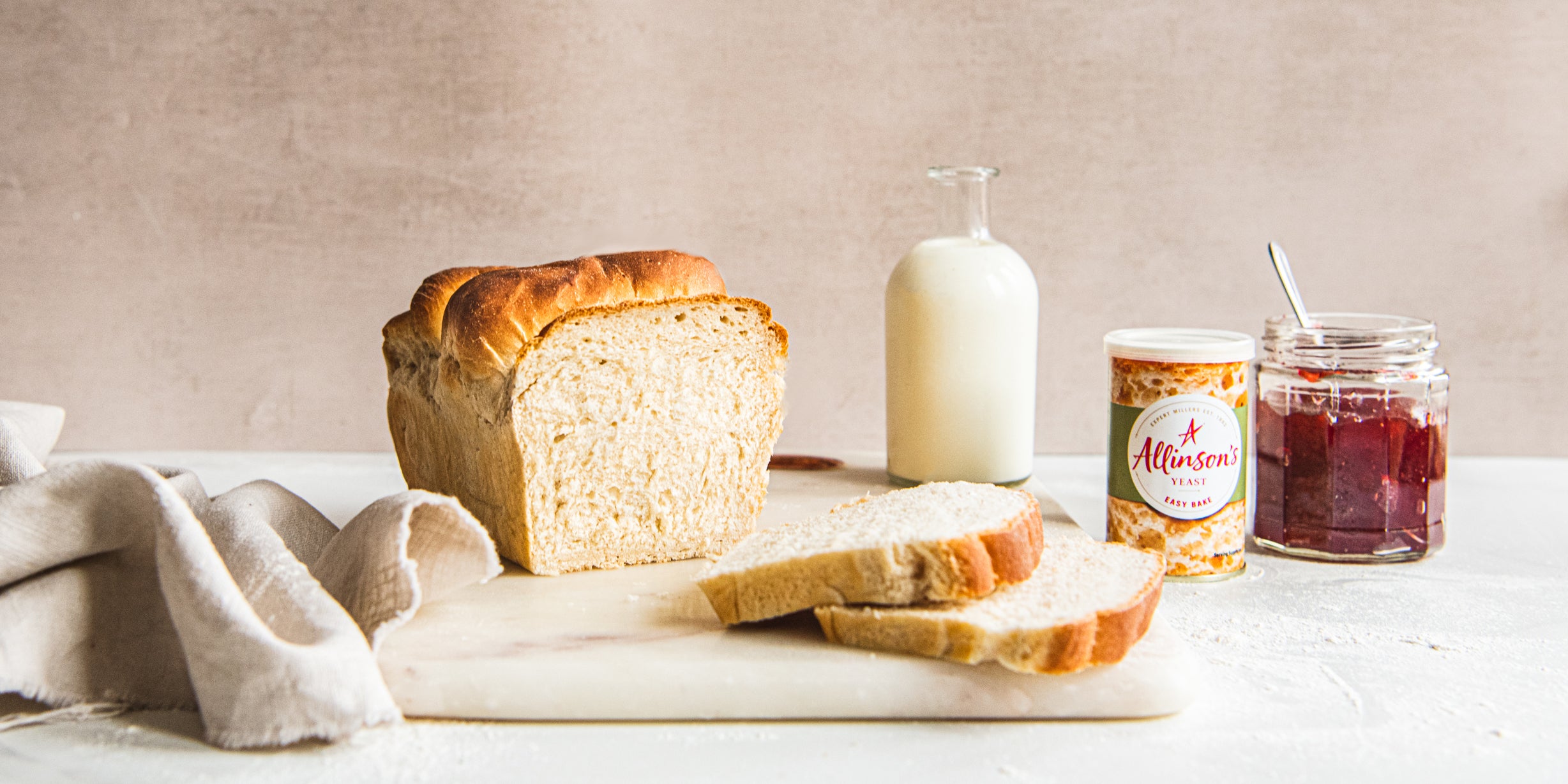 Shokupan Milk Bread sliced on a serving board, next to a glass bottle of milk, and jar of jam and a tin of Allinson's Yeast