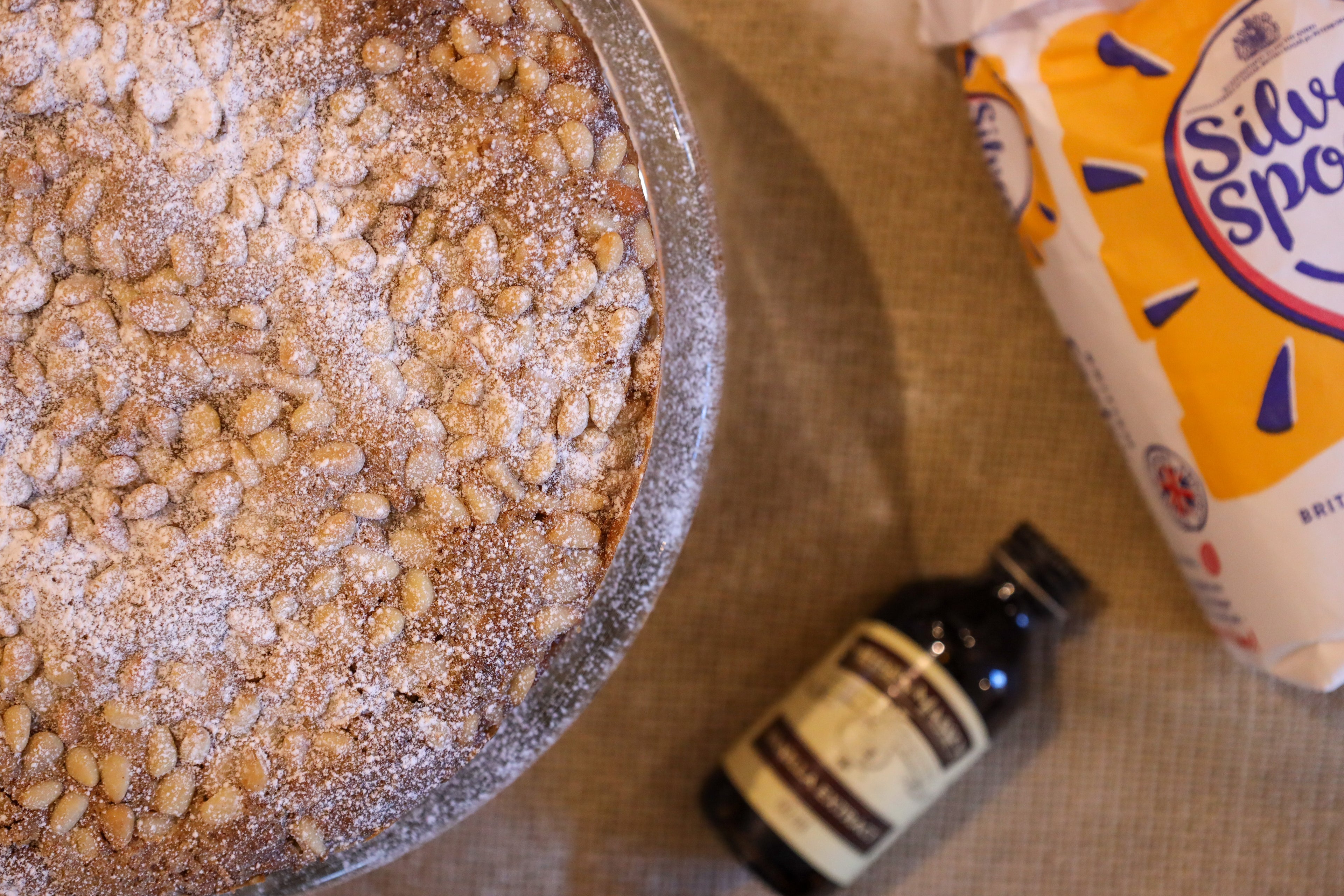 Top down image of apple cake with pine nuts, vanilla bottle and sugar packaging