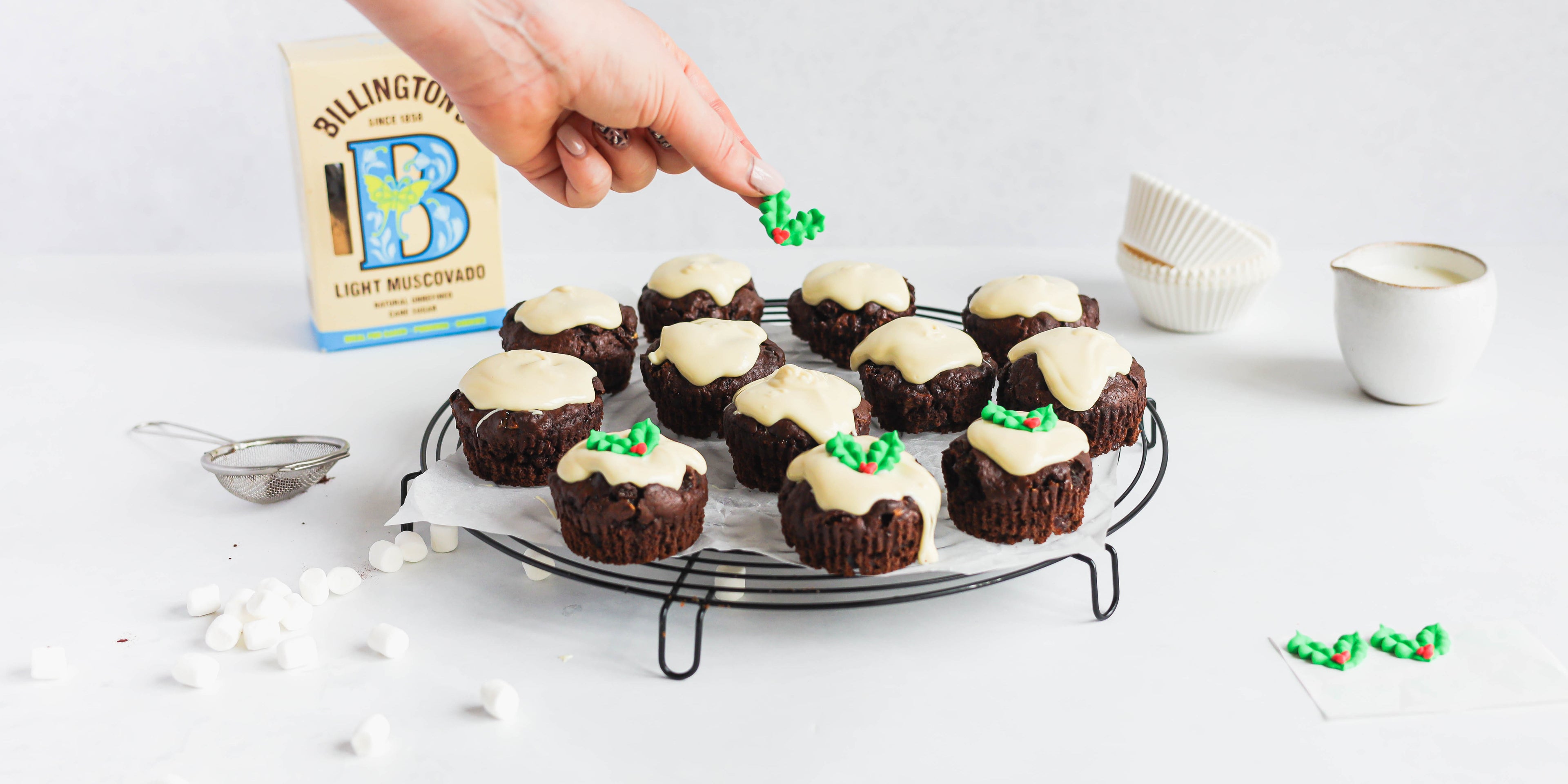 Mini Rocky Road Christmas Pudding Muffins on a wire rack, being hand decorated with a box of Billington's Light Muscovado in the background