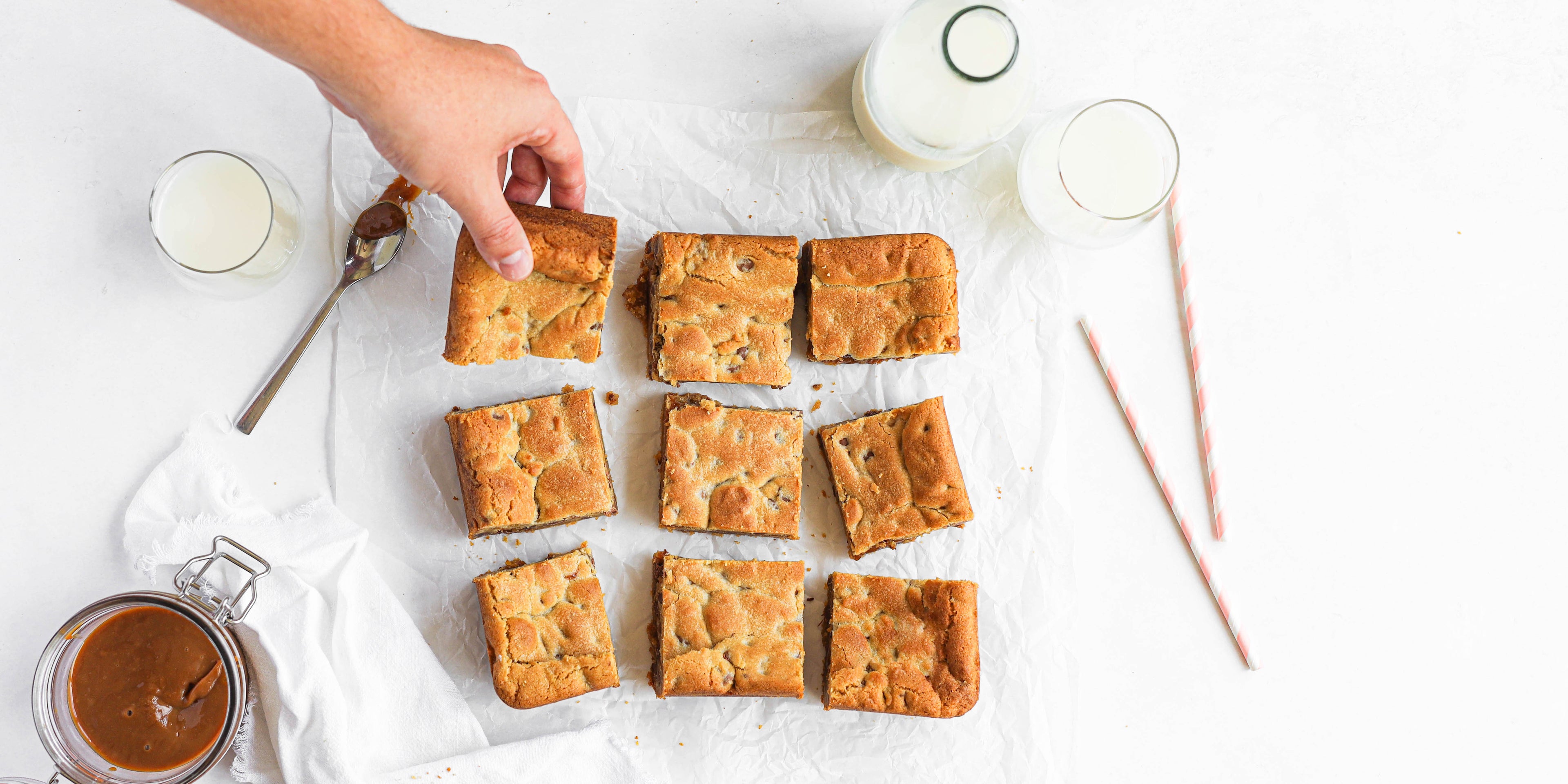 Salted Caramel Cookie Traybake cut onto baking paper, with hand reaching for a square 