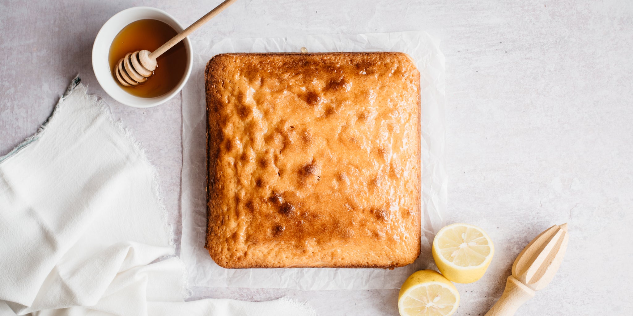 Uncut square lemon drizzle cake with a small bowl of honey and lemons beside it