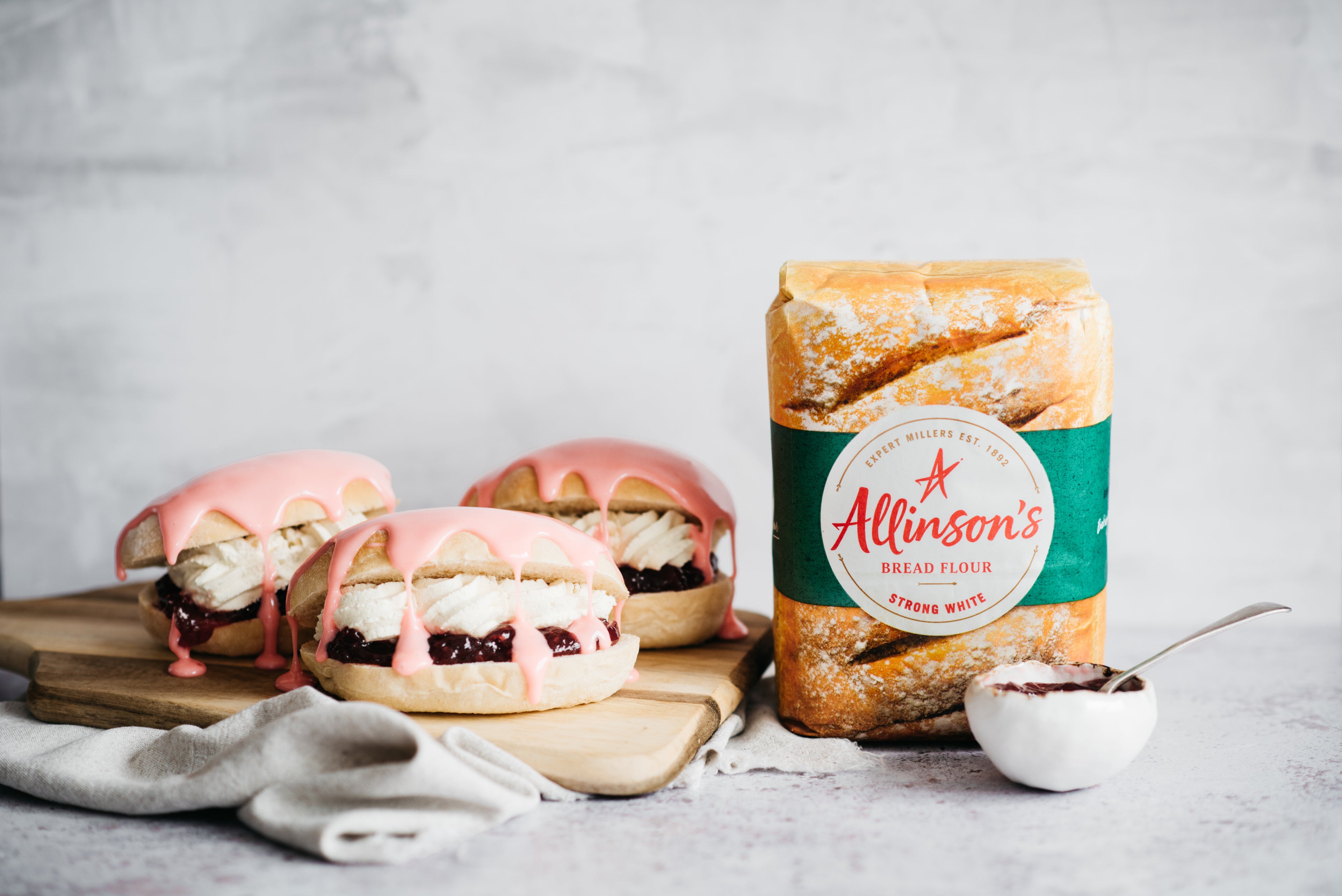 Iced Finger Buns on a wooden serving board, drizzled in pink icing next to a bag of Allinson's strong white flour and a bowl of jam