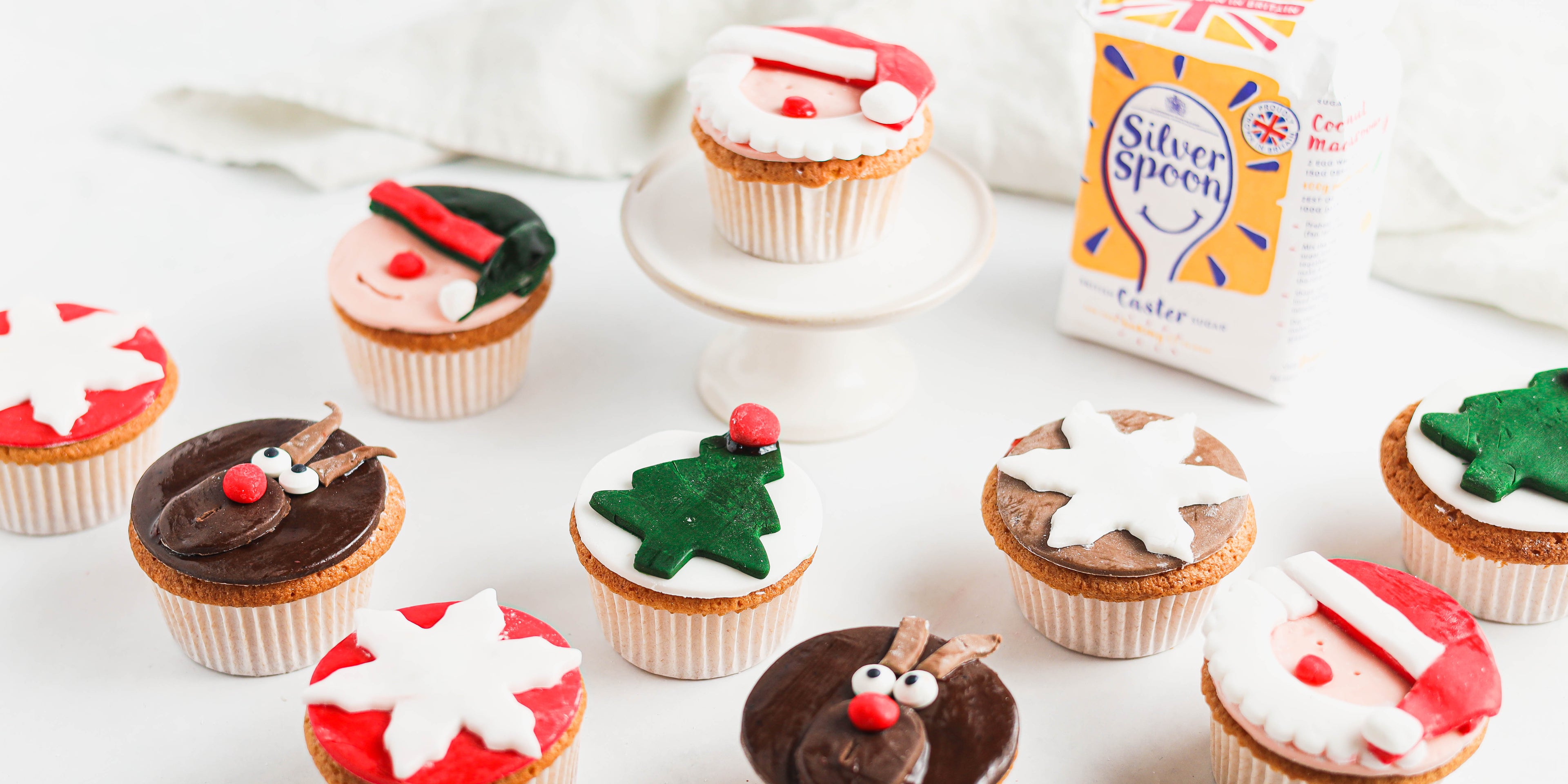 Close up of Christmas Cupcakes decorated with reindeer, stars, trees and santa next to a bag of silver spoon caster sugar