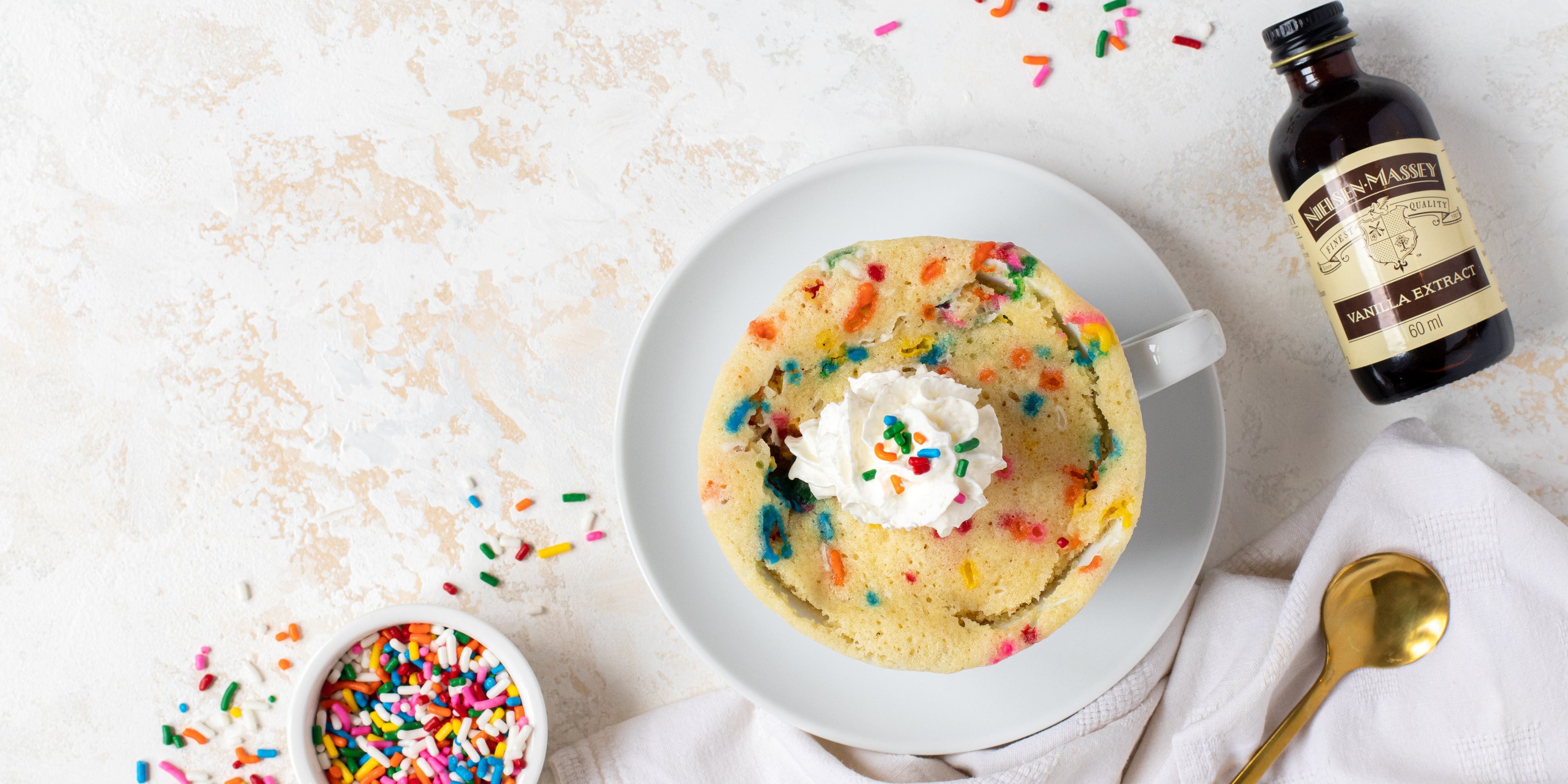 Top view of Birthday Cake Mug Cake with sprinkles on top, next to a bottle of Nielsen-Massey Vanilla extract