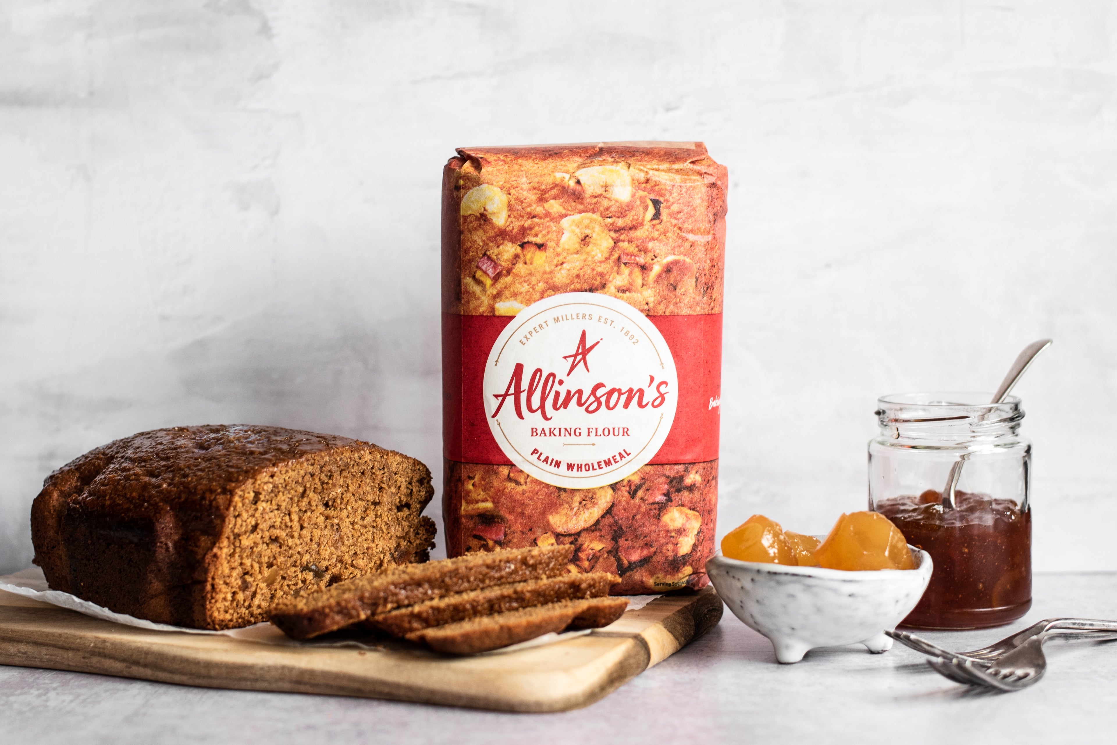 Gingerbread loaf with slices infront, flour pack, bowl of ginger and marmalade in a jar