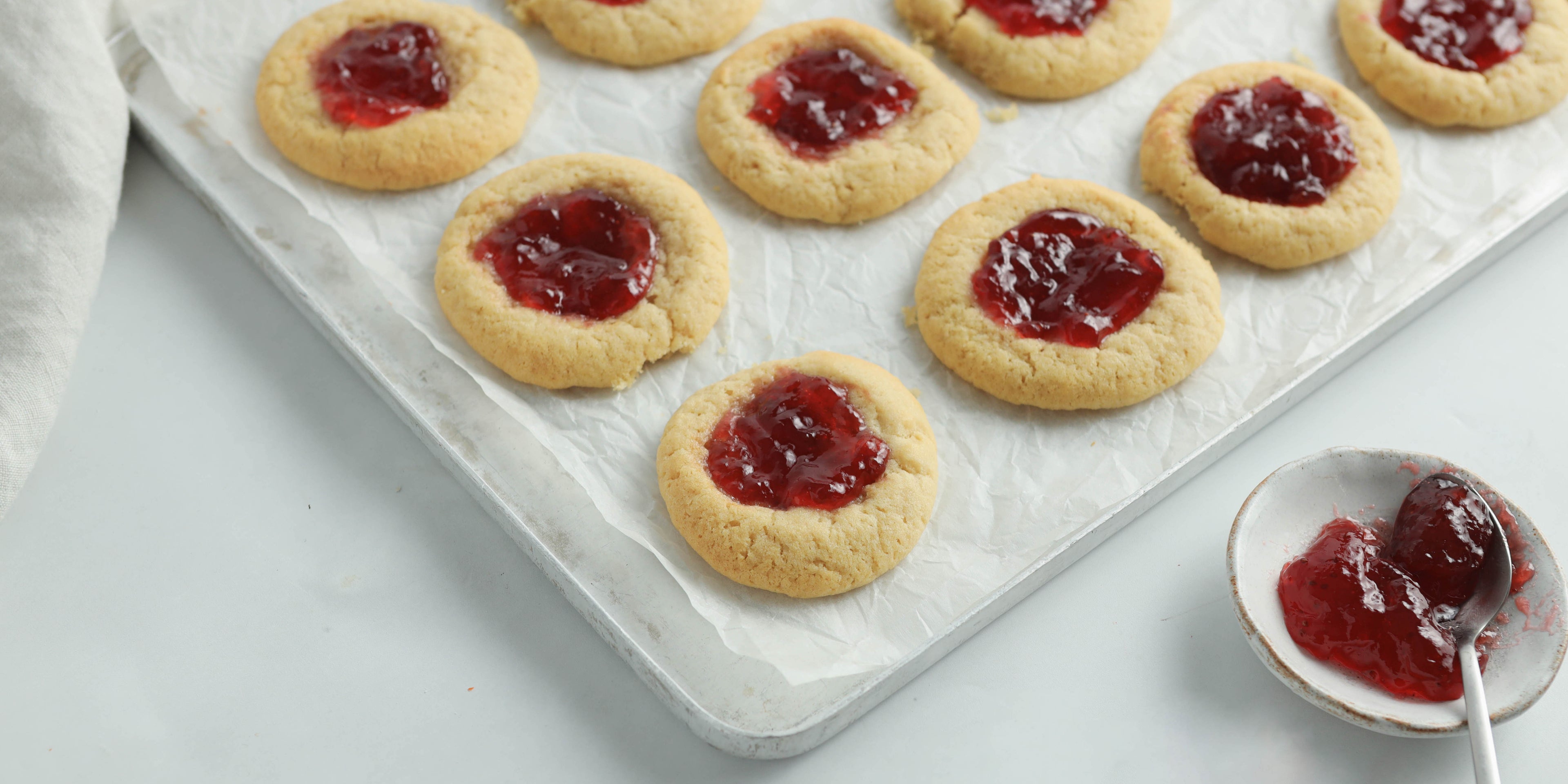 Close up shot of jammy biscuits on a baking tray with a spoon in a small pot of jam