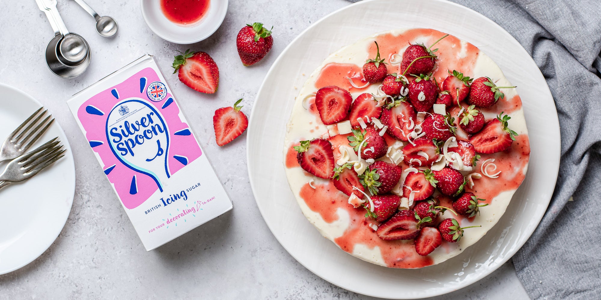 White Chocolate & Strawberry Cheesecake from above view, with slices of strawberries lay next to a box of Silver Spoon Icing sugar