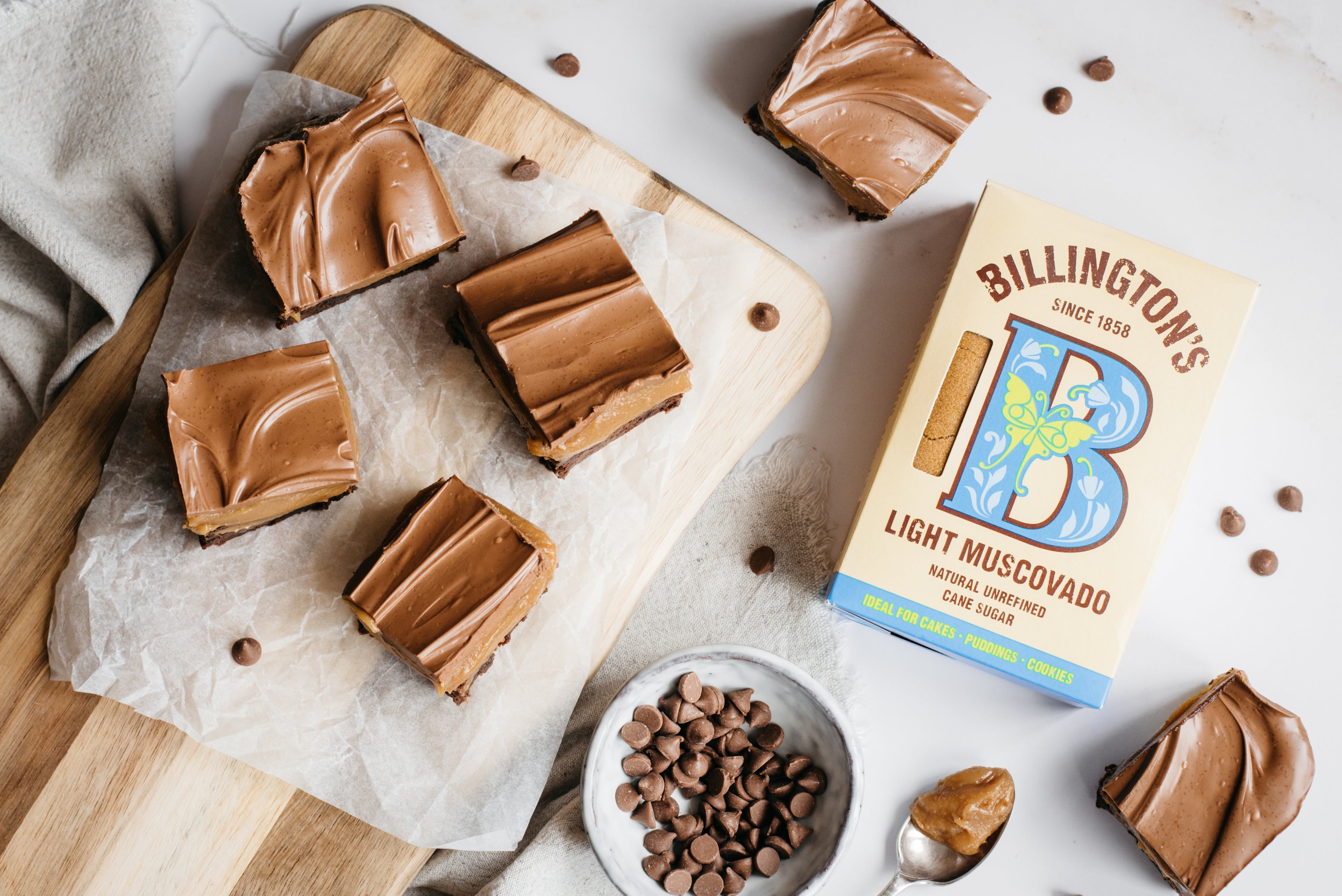 Flat lay of Brownie Based Millionaires Traybake with Billington's light muscovado sugar, a bowl of chocolate chips and a spoonful of caramel