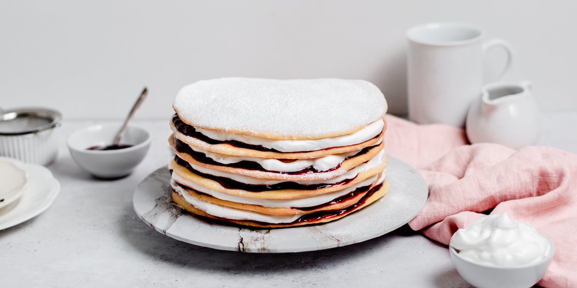 Danish Layer Cake dusted in icing sugar, served on a marble plate, with a pink linen cloth in the foreground and a bowl of whipped cream in the foreground