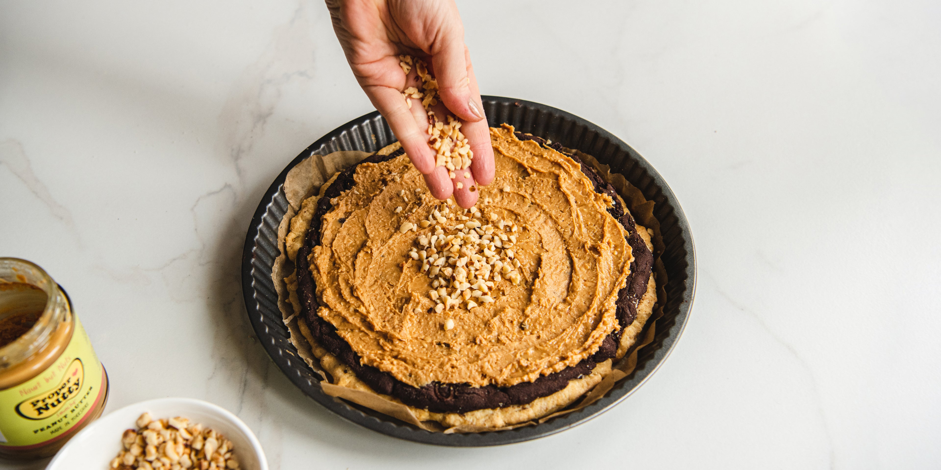 Easter Mazurek being hand sprinkled with peanuts, on top of a peanut layer next to a jar of Proper Nutty