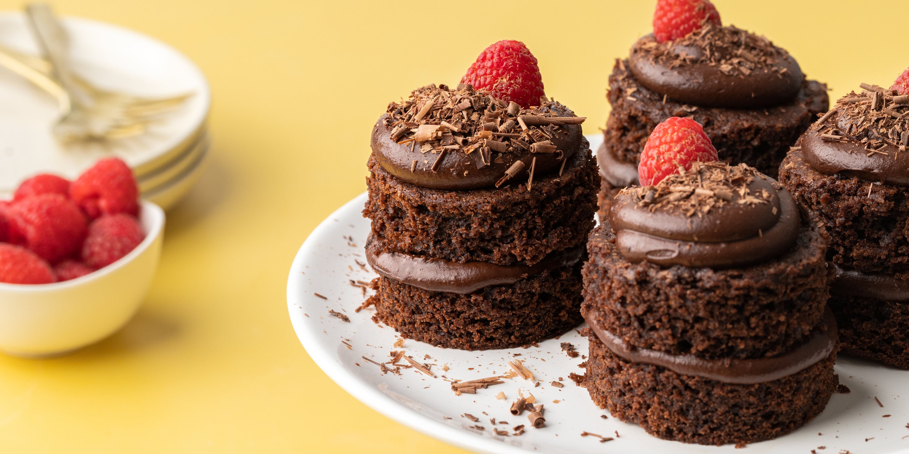 4 mini chocolate cakes on a plate with raspberries on top, beside a white bowl of raspberries