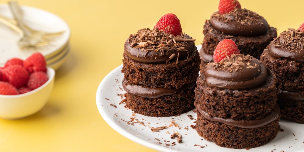4 mini chocolate cakes on a plate with raspberries on top, beside a white bowl of raspberries