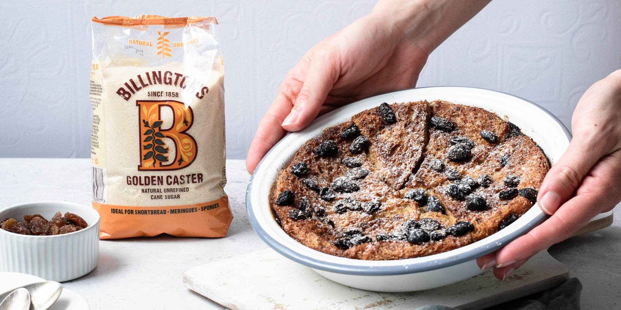 Classic Bread & Butter Pudding in white bowl being held by hands. Sugar pack and raisins in bowl